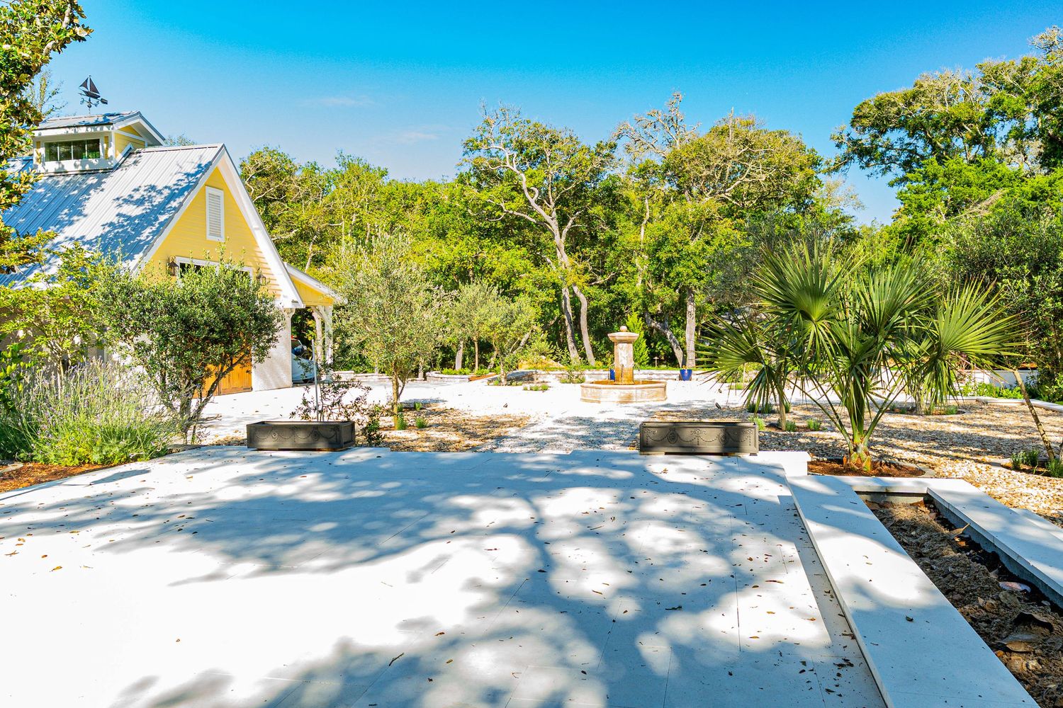 White concrete patio surrounded by palm trees and lush greenery beside a charming yellow cottage on a sunny day.