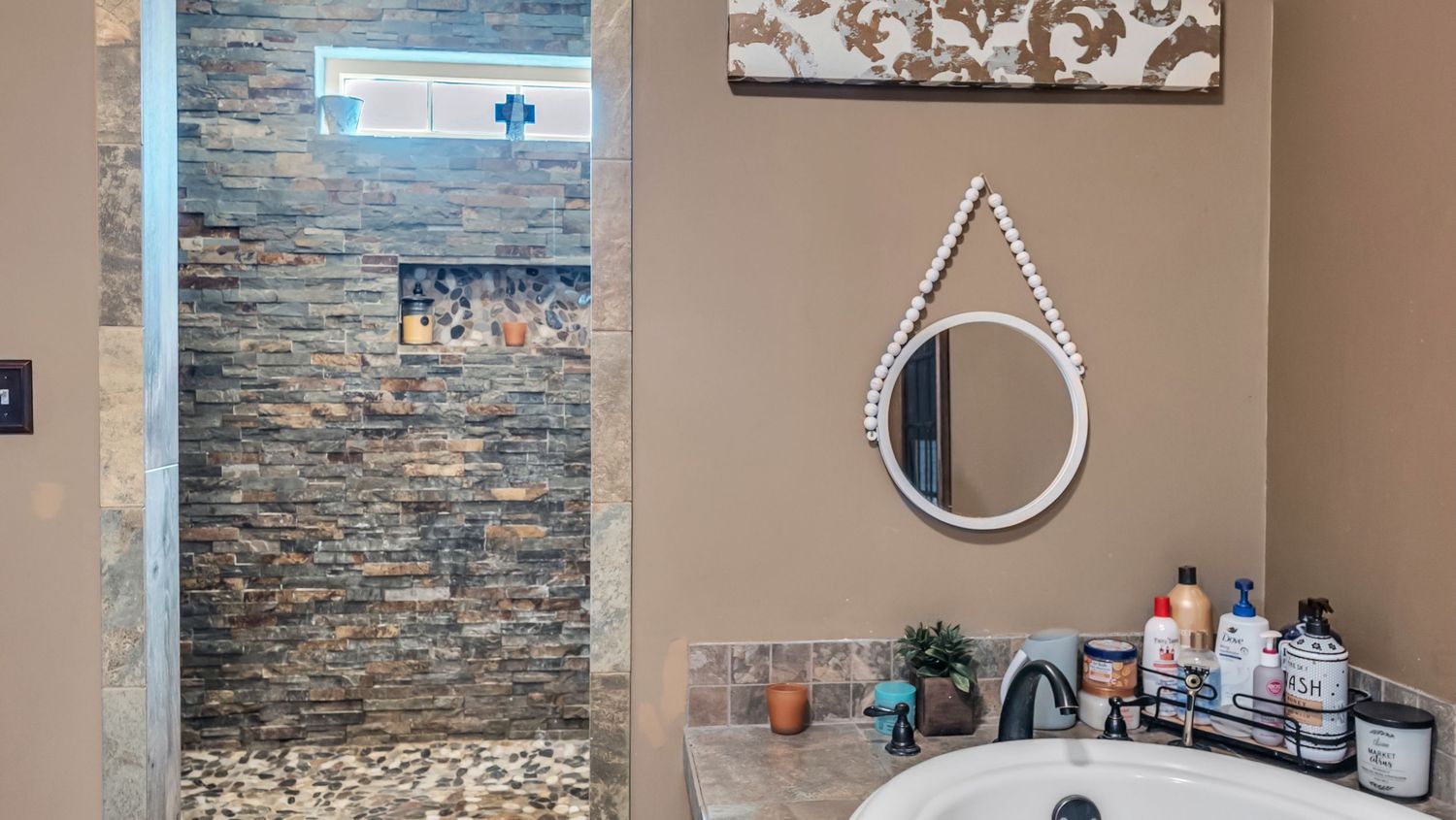 Luxury bathroom featuring a stone-tiled walk-in shower, round mirror, and marble sink with toiletries on the counter.