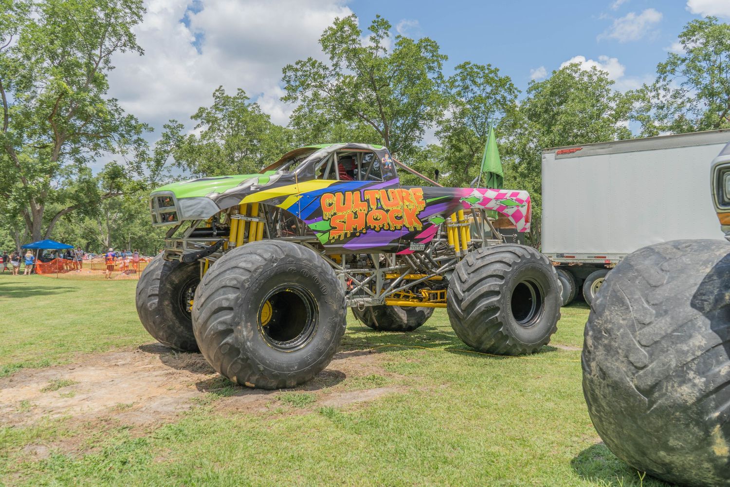 Colorful monster truck with massive tires on green grass at an outdoor event under a partly cloudy sky.
