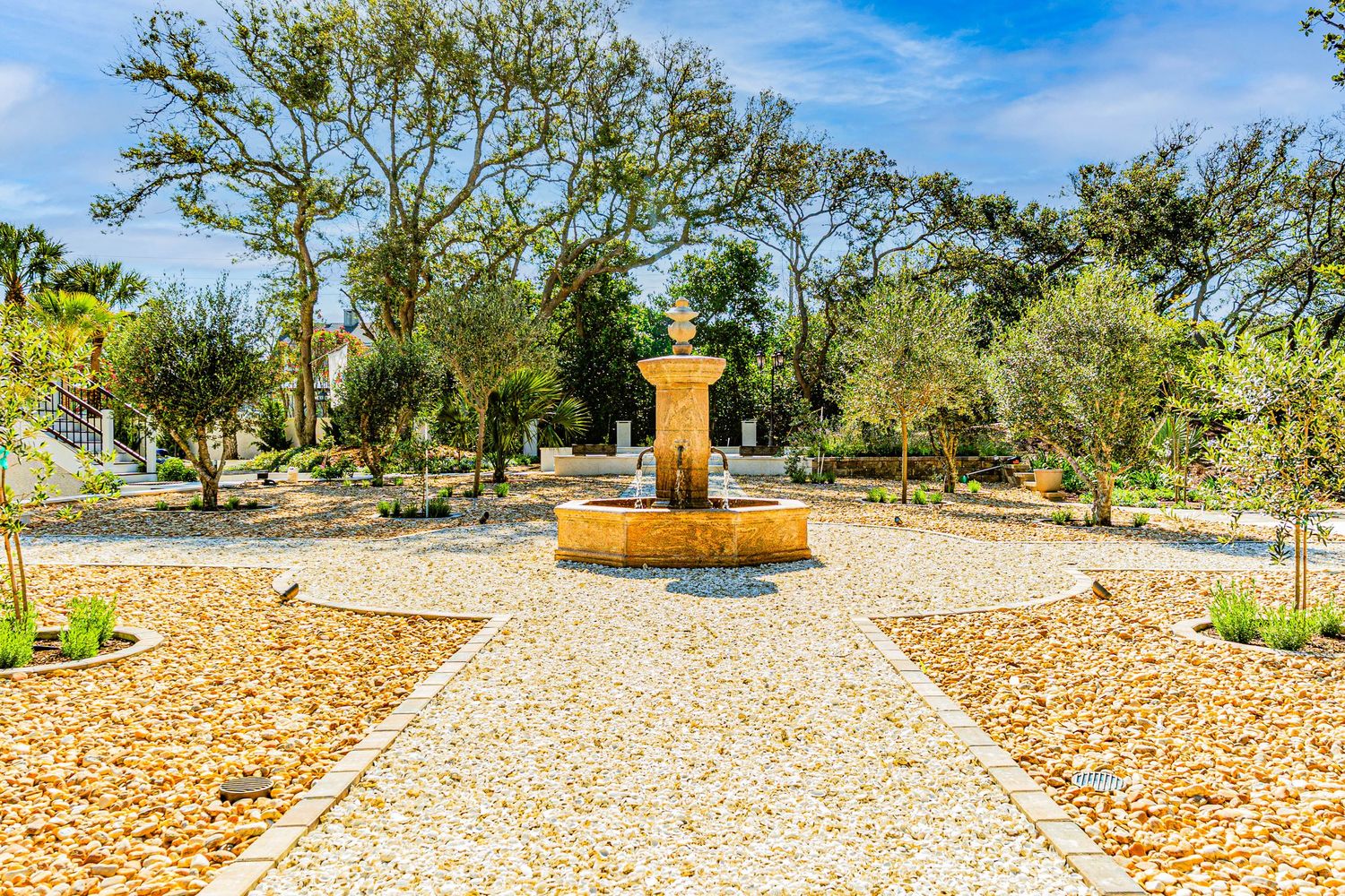 Sunny garden with a stone fountain centerpiece, gravel pathways, and lush green trees in a serene outdoor setting.