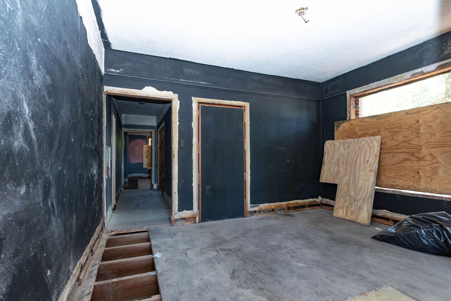 Unfinished room with dark walls, exposed wood flooring, doorframes, and plywood sheets during home renovation.