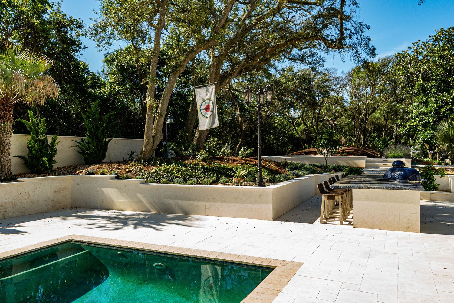 Outdoor pool area with travertine patio, raised garden beds, lush trees, and a decorative flag on a sunny day.