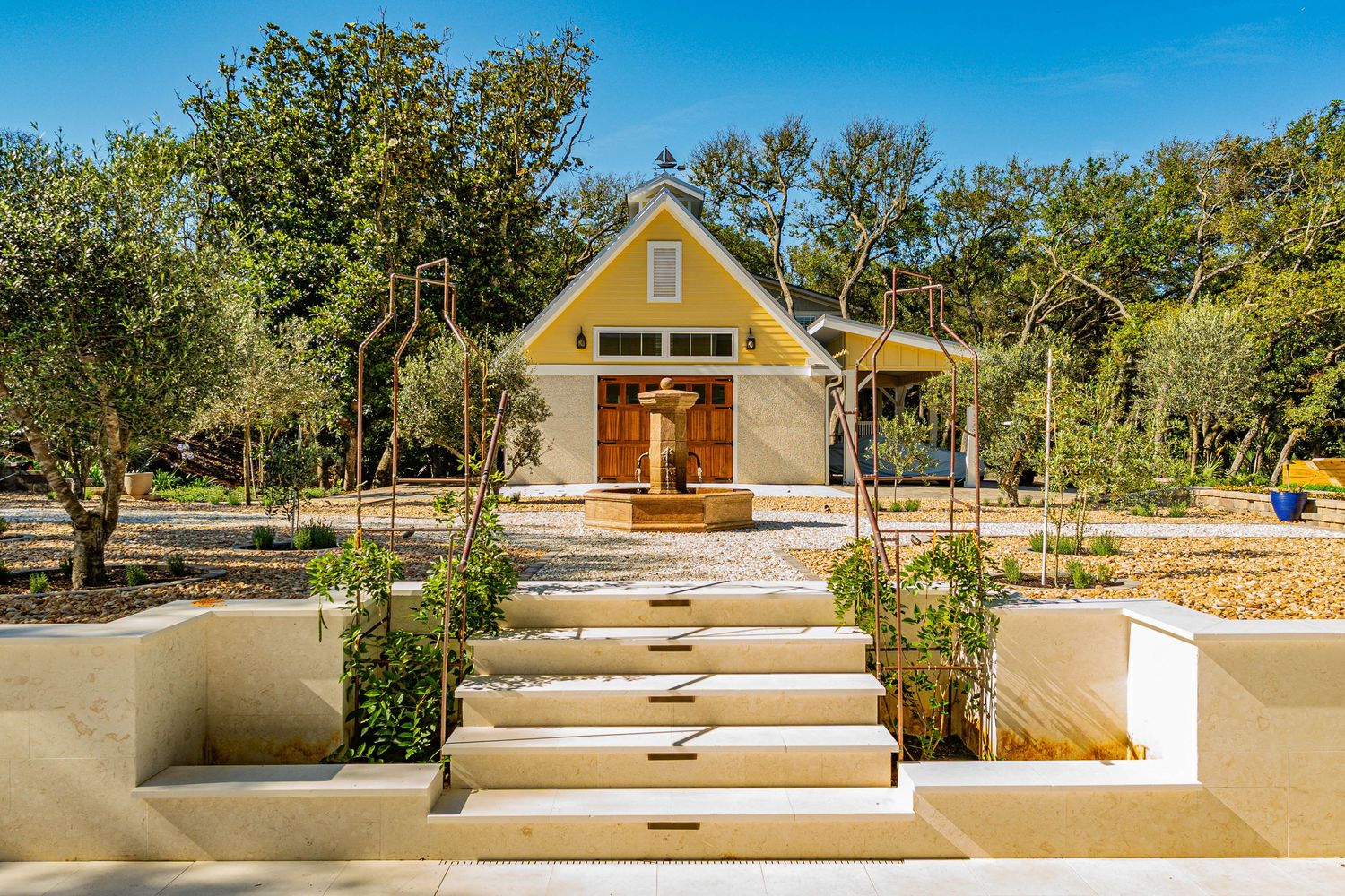 Yellow cottage with gabled roof surrounded by trees, featuring stone steps and landscaped garden in warm sunlight.