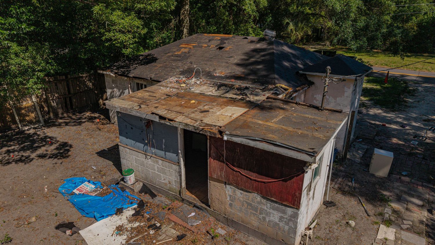 Aerial view of a dilapidated shed with damaged roof, debris, and blue tarp on the ground surrounded by trees.