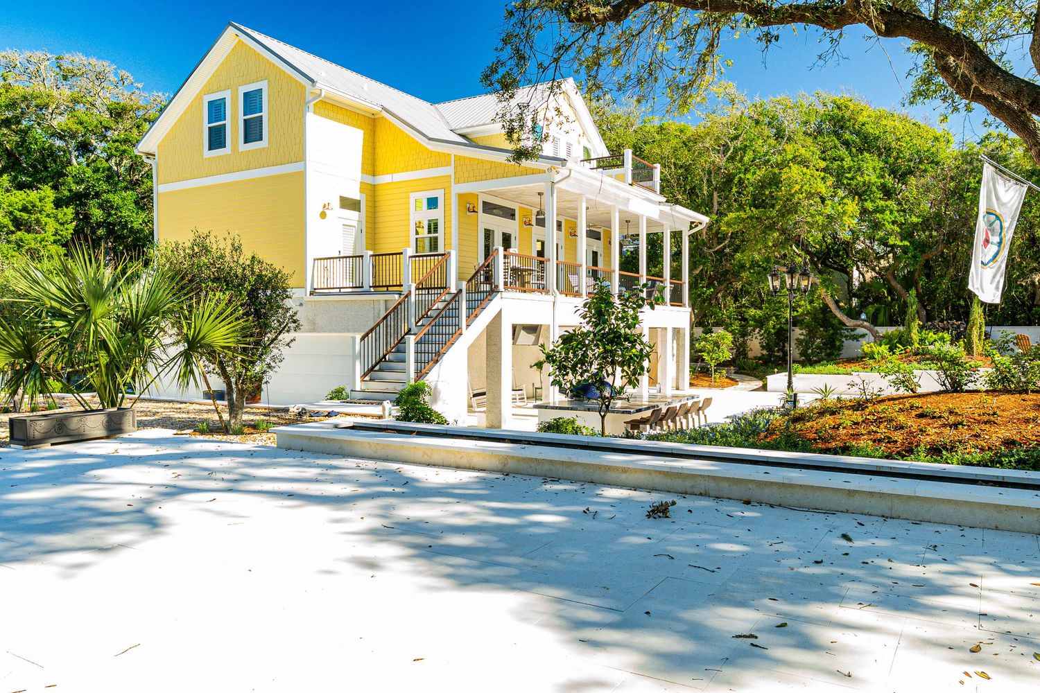 Yellow two-story coastal home with wrap-around porch, grand staircase, and lush tropical landscaping on a sunny day.