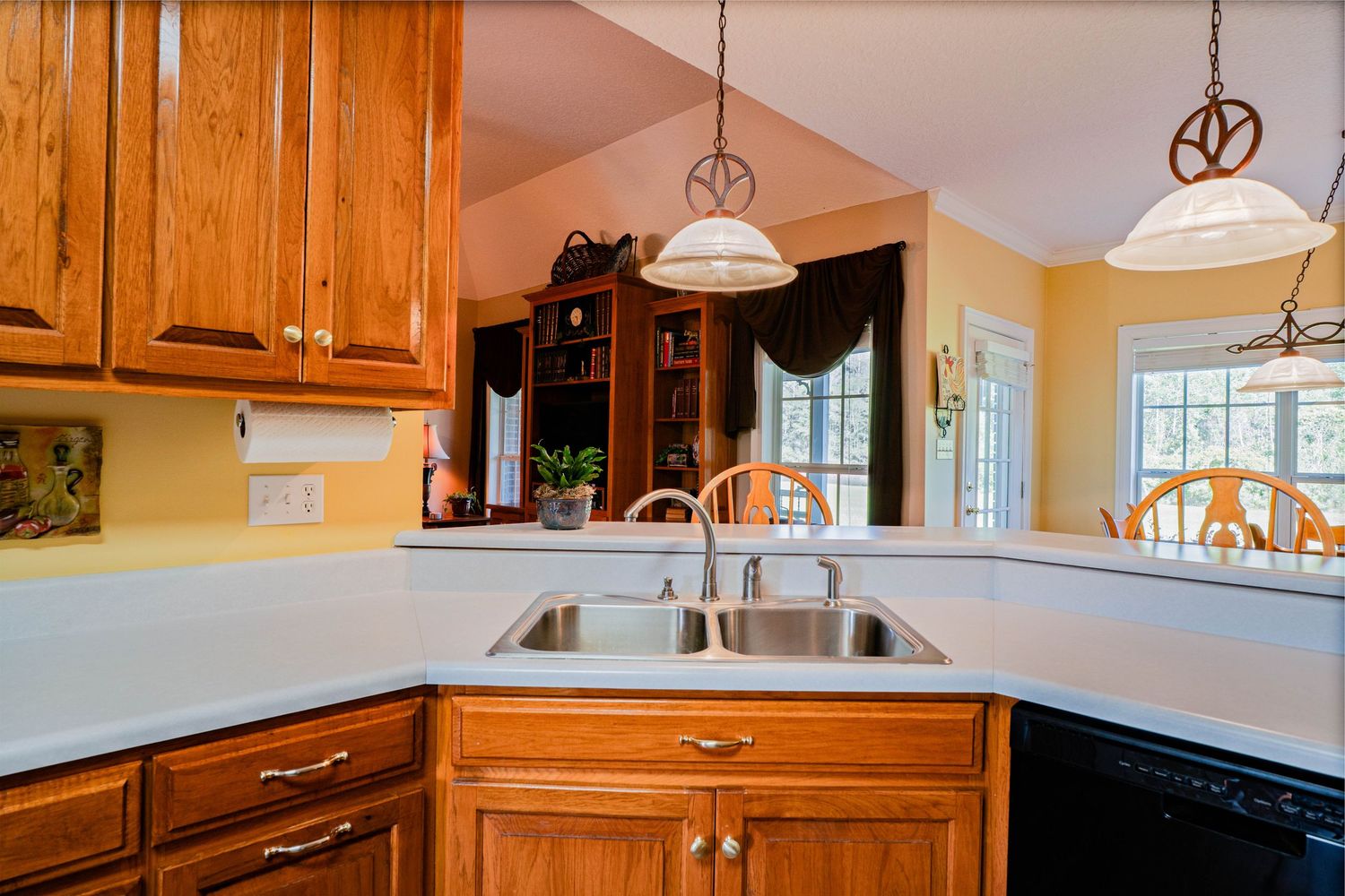 Bright kitchen with oak cabinets, double stainless steel sink, white countertops, and pendant lights over a open floor plan.