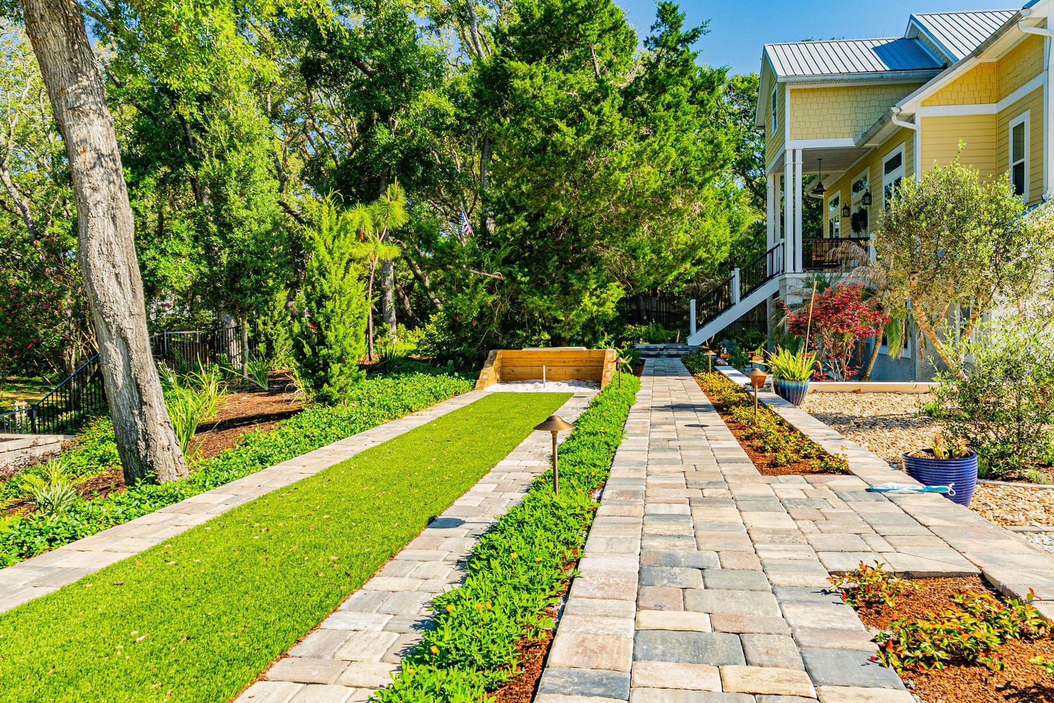 Lush backyard garden with stone paver walkway, manicured green lawn, and tropical trees leading to a yellow house.