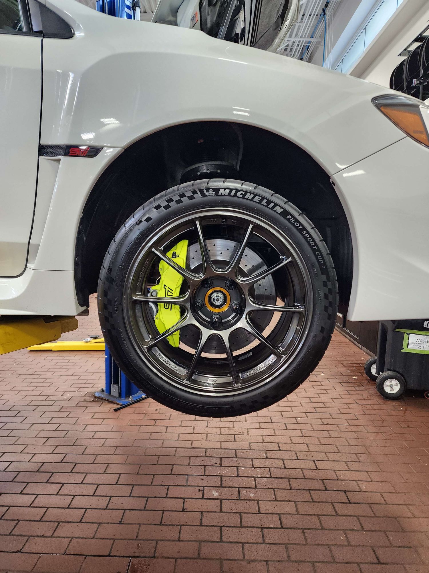 White car with Michelin tires on dark multi-spoke wheels with yellow brake calipers on lift.