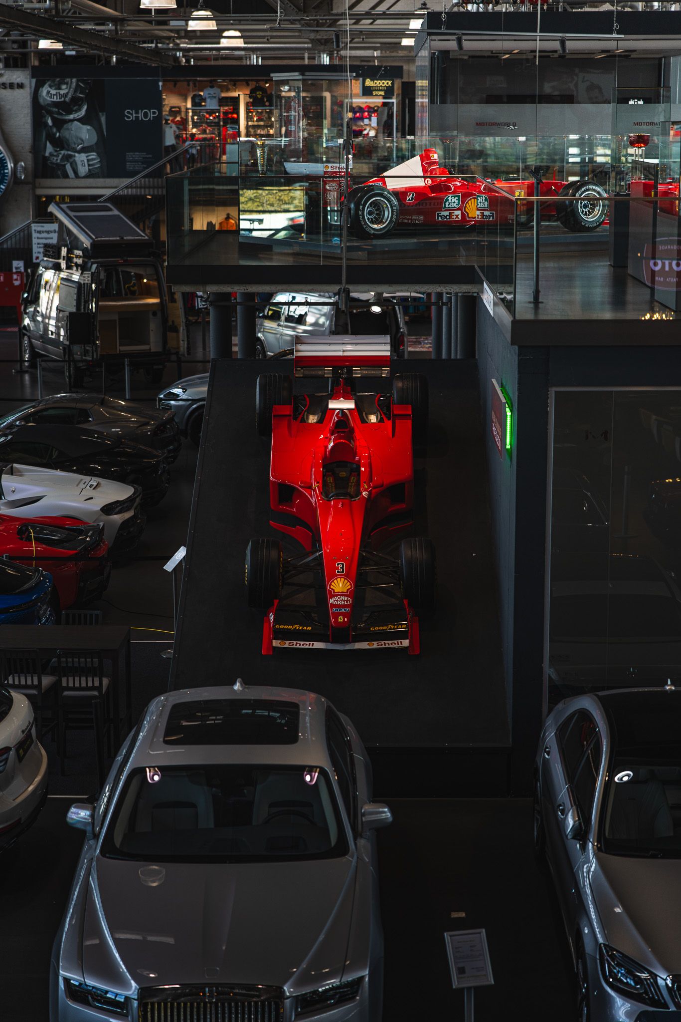 Red Ferrari Formula 1 car displayed on a pedestal surrounded by luxury cars indoors.