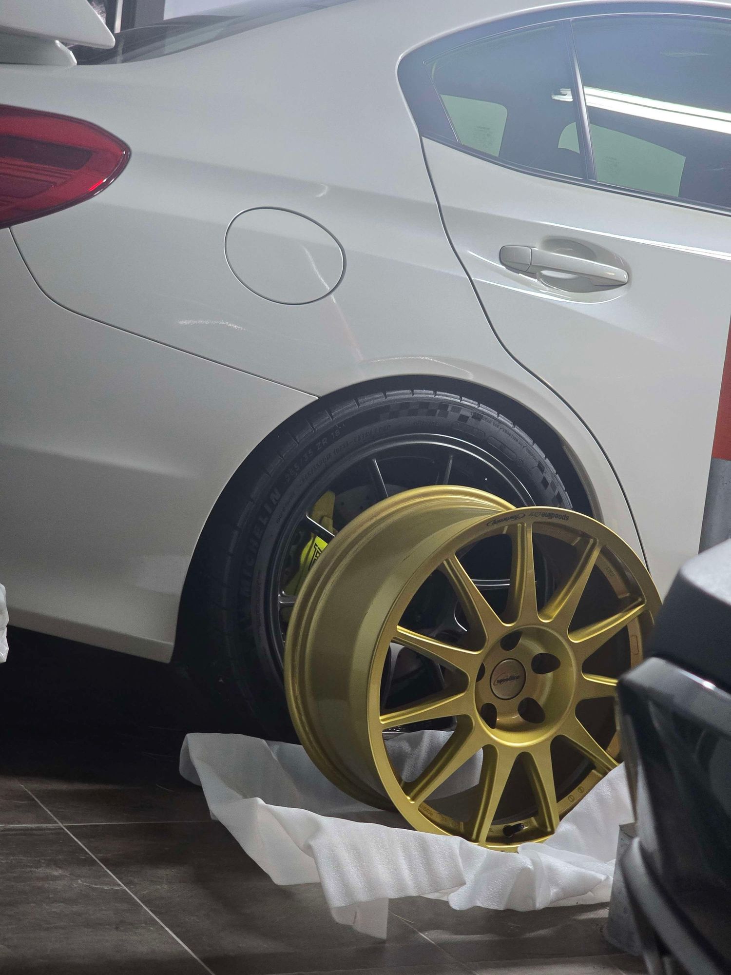 Gold aftermarket alloy wheel leaning against white car in automotive shop.