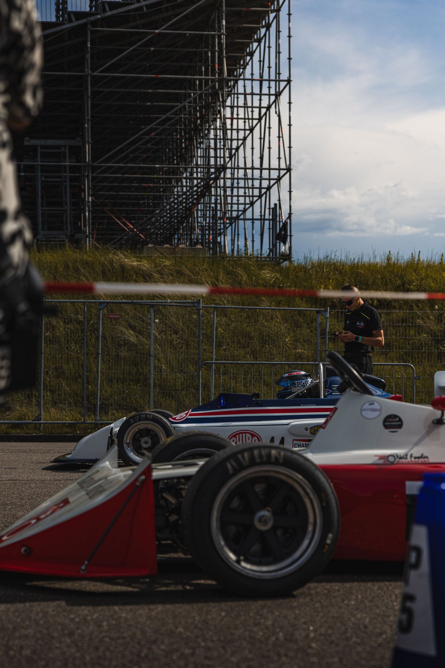 Red and white Formula racing car on track with grandstand scaffolding and official in background.