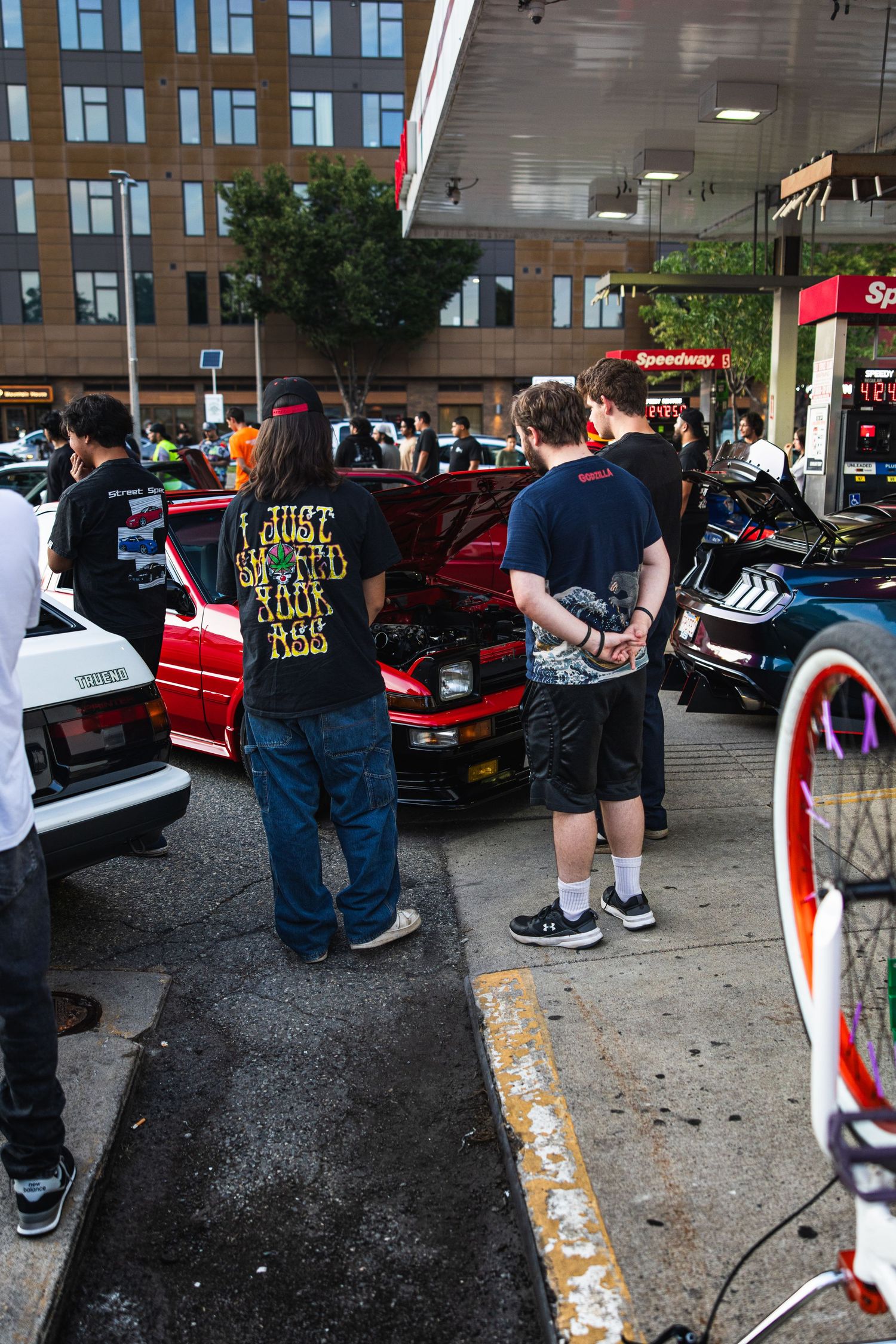 Car enthusiasts gather at a Speedway gas station car meet, admiring a red car with hood open.