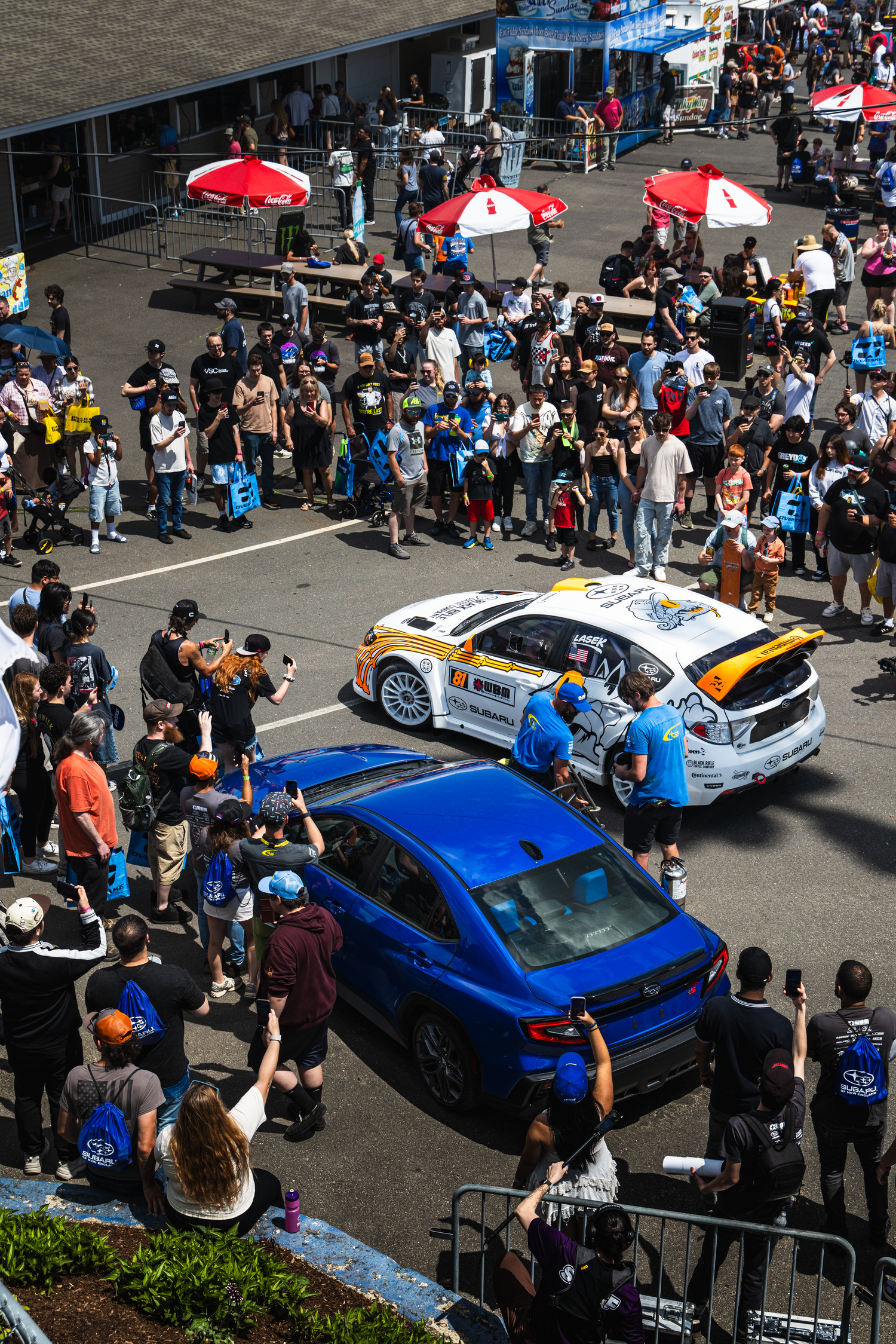 Aerial view of rally cars surrounded by crowds at an outdoor motorsport event on a sunny day.