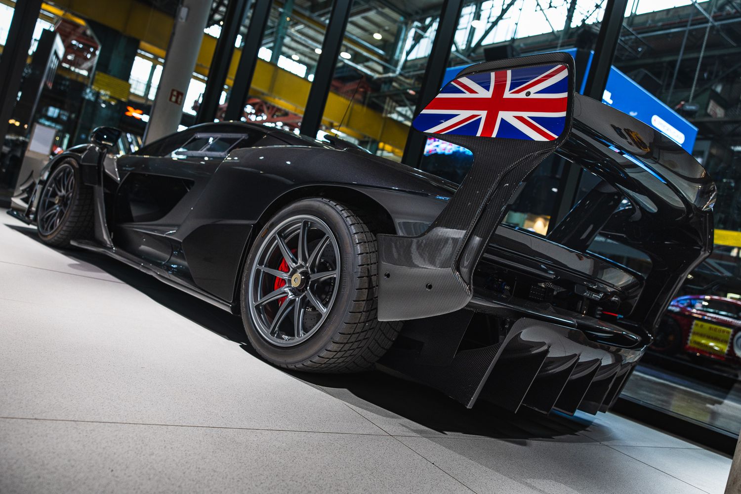Sleek black McLaren supercar with Union Jack mirror displayed indoors under bright exhibition lighting.
