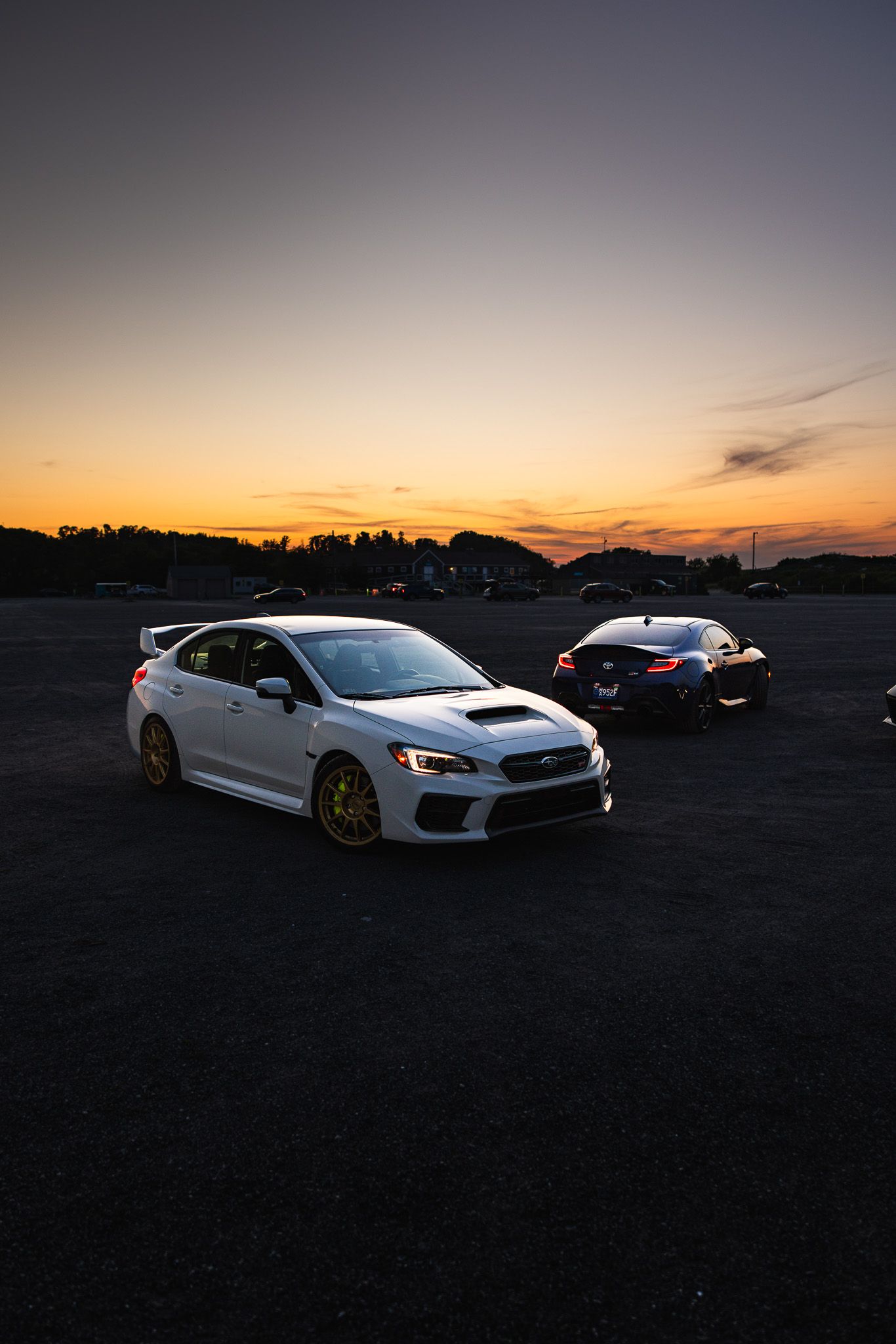White Subaru WRX STI with gold wheels parked at sunset alongside a dark sports car.