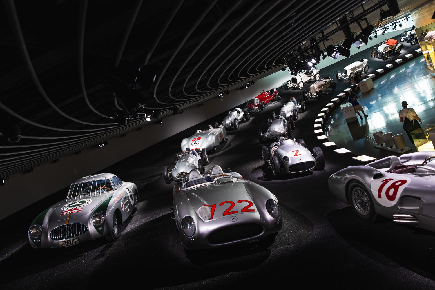 Vintage silver Mercedes-Benz racing cars displayed in a dramatic curved exhibition hall at a classic car museum.