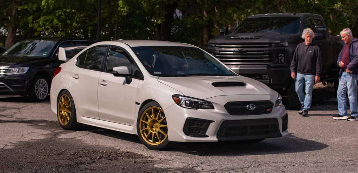 White Subaru WRX STI with gold wheels parked at an outdoor automotive meet on pavement.