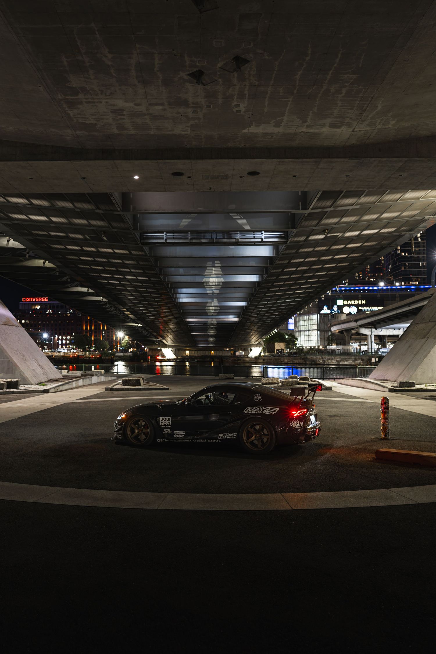 Dark sports car with rear spoiler parked under illuminated urban overpass at night with city lights reflecting.