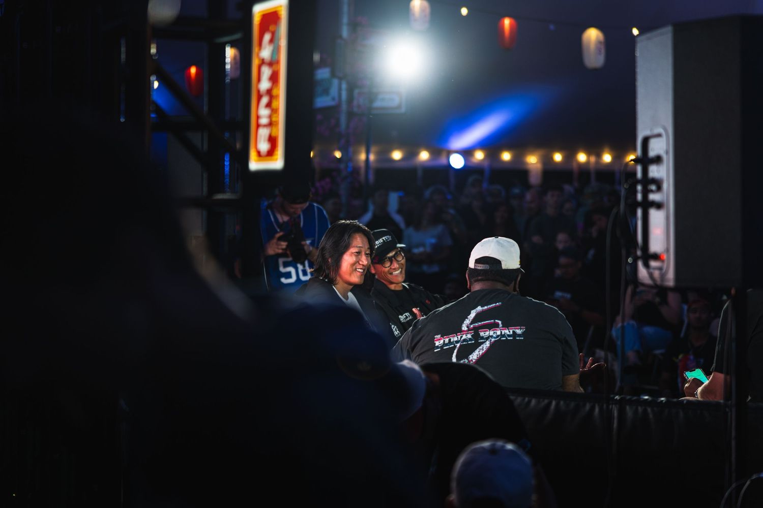 Crowd gathered at a dimly lit indoor event venue with stage lighting and performers visible in the foreground.