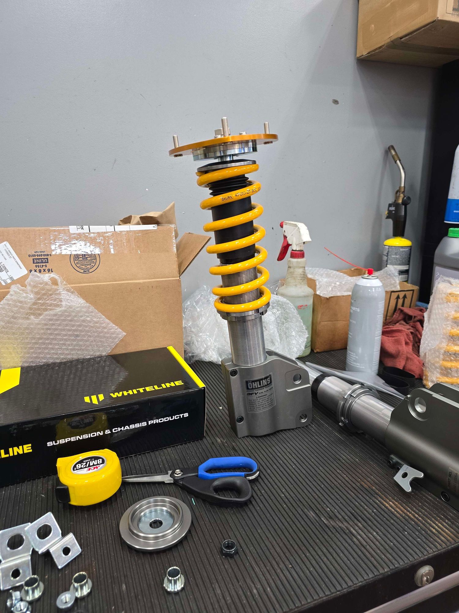 Yellow Ohlins coilover suspension assembly on workbench with Whiteline parts and tools in automotive garage.