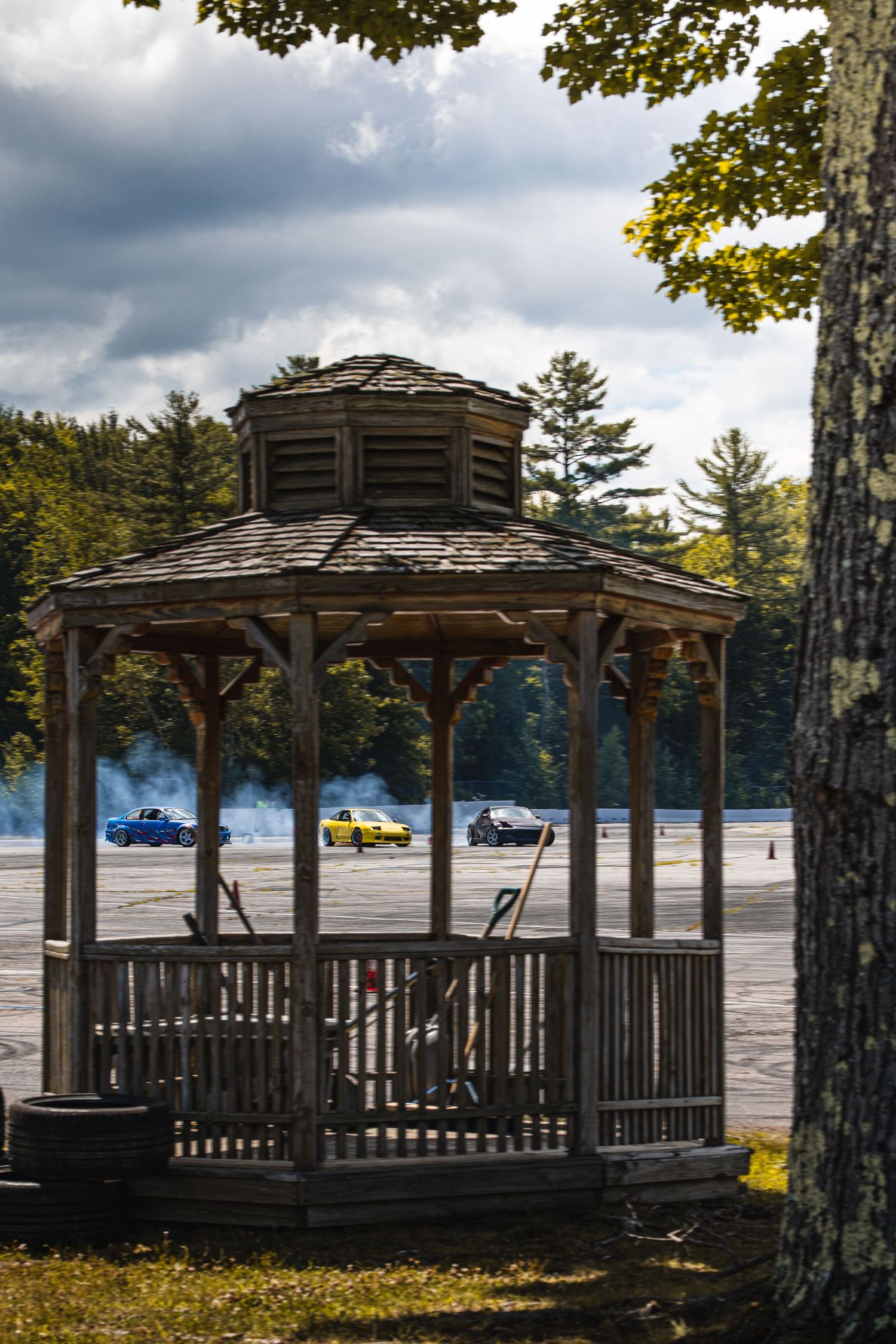 Wooden gazebo in foreground with racing cars on a track behind it, surrounded by trees under a cloudy sky.