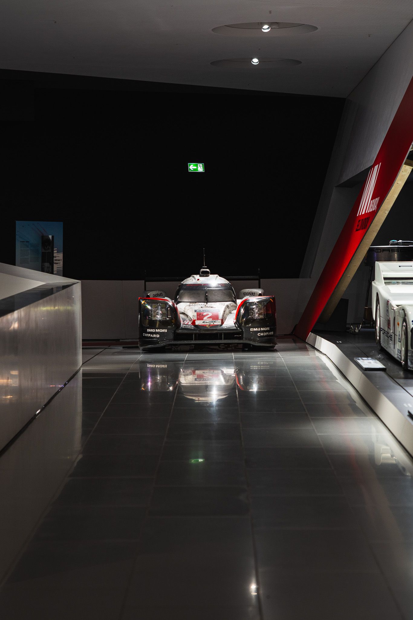 Porsche LMP1 hybrid racing car displayed in a dark dramatic museum corridor with reflective floor.