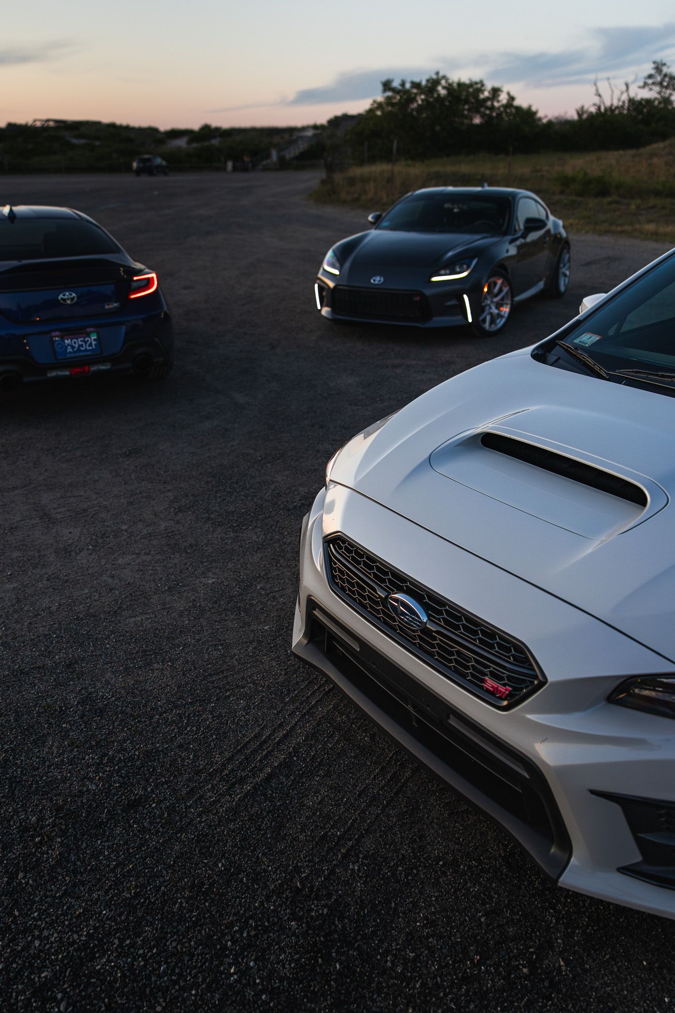 Three sports cars including a white Subaru WRX STI and Toyota GR86 gathered at dusk on open pavement.
