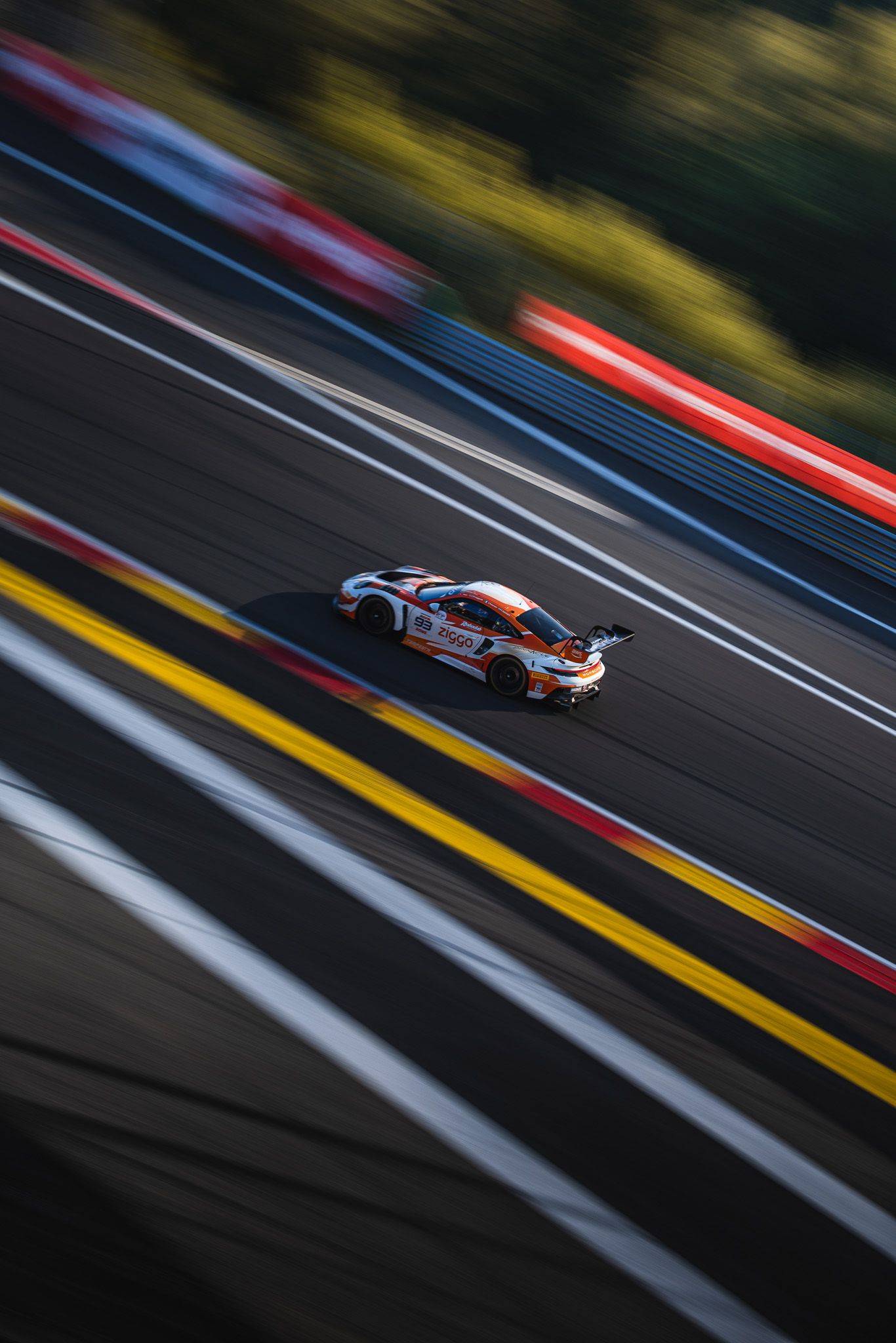 Aerial view of a white racing car speeding on a sunlit track with colorful barriers at a motorsport circuit.