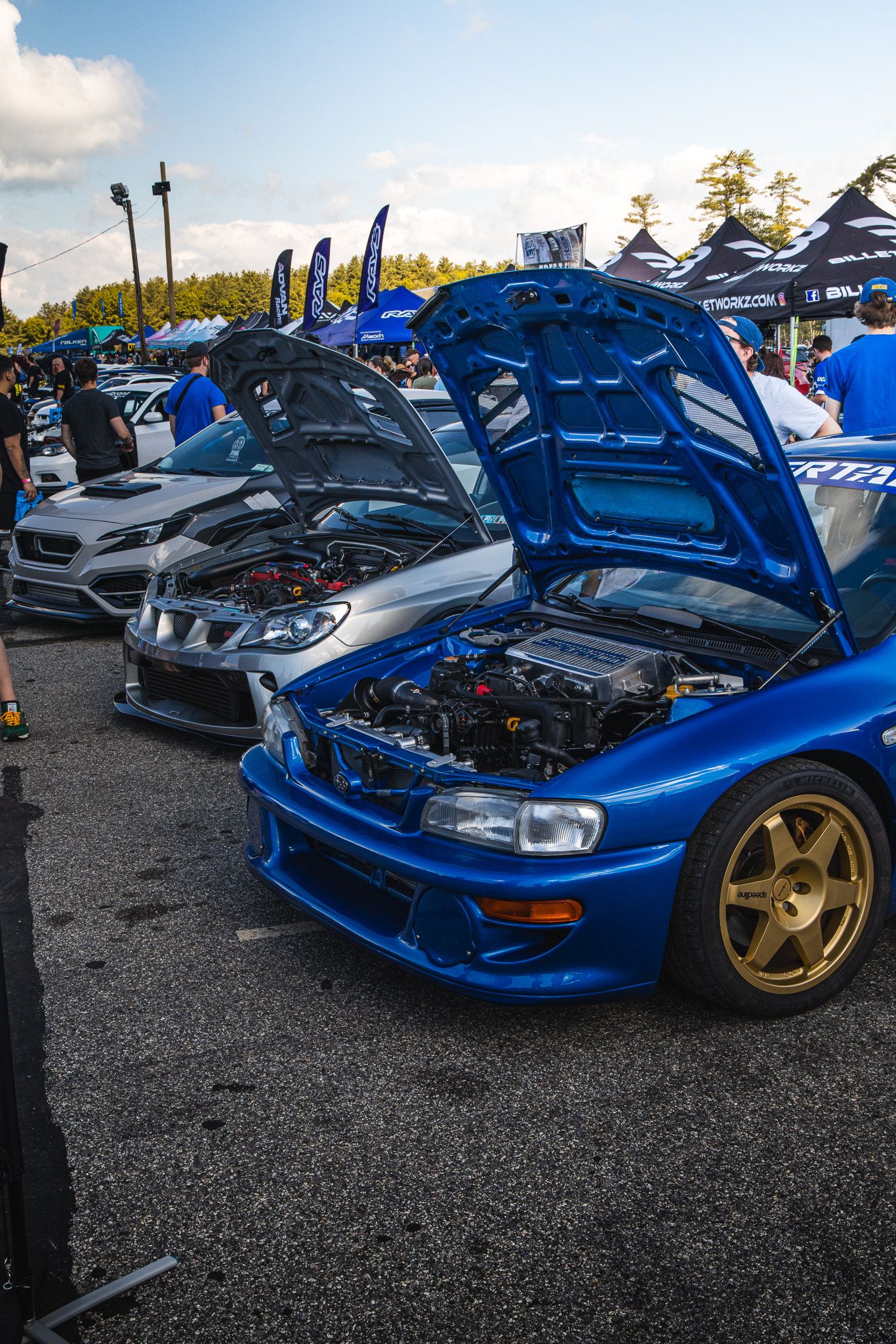 Blue Subaru Impreza GC8 with gold wheels and open hood at a car show event.