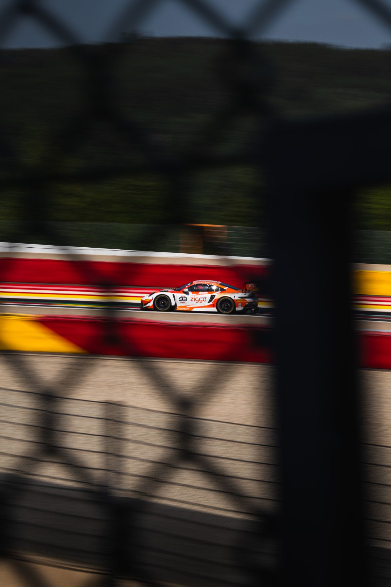 White GT race car captured through fence at speed on Spa-Francorchamps circuit in golden light.