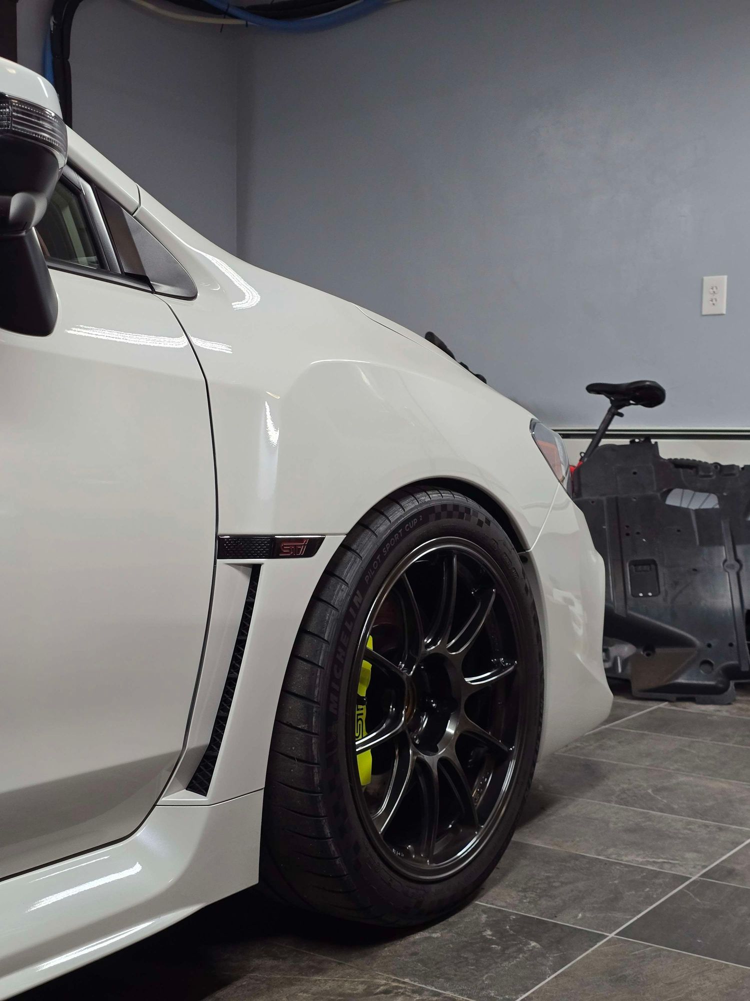 White Subaru WRX STI with black wheels and yellow brake calipers in a clean garage setting.