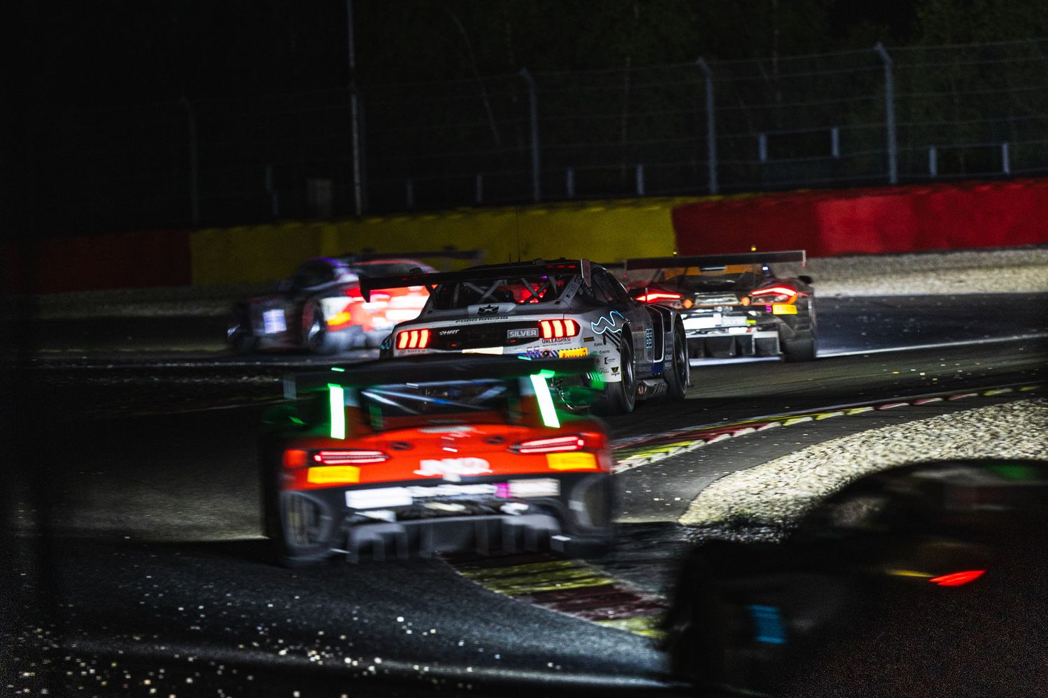 GT racing cars battle in wet night conditions on a circuit, with rain-soaked track reflecting colorful lights.