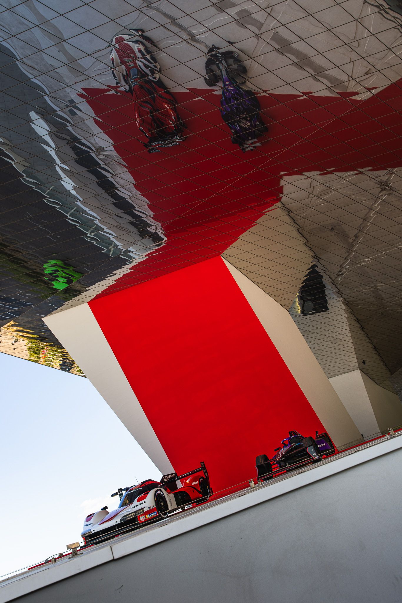 A racing car reflects dramatically in a mirrored architectural ceiling beside a bold red structure at a motorsport venue.
