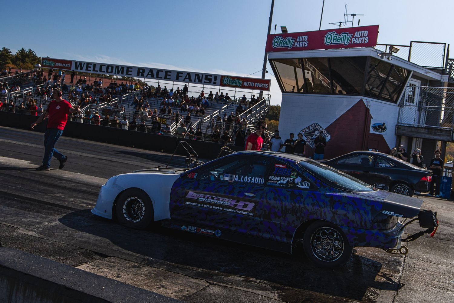 Two drag racing cars compete on a track with a crowded grandstand and Welcome Racers sign in the background.
