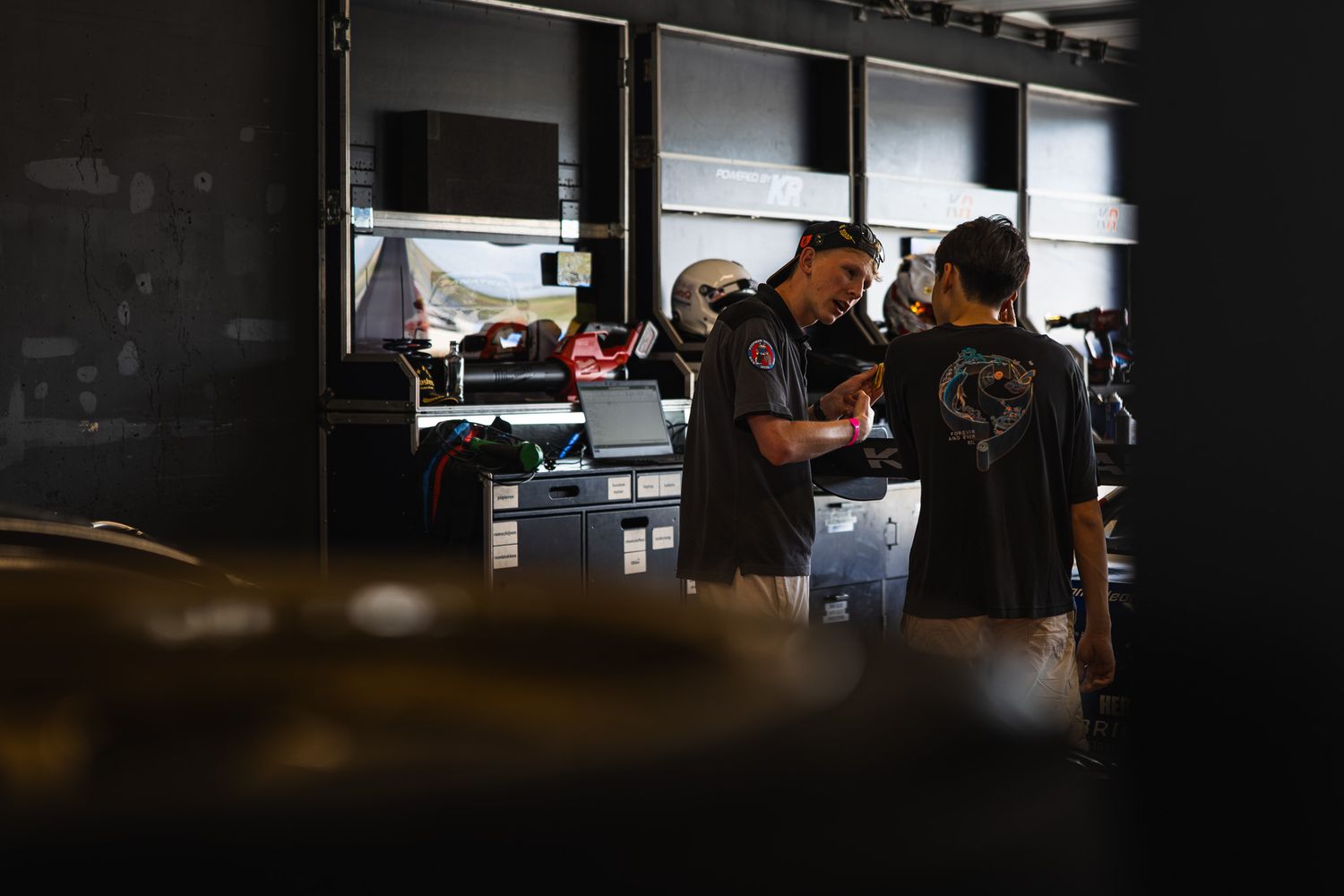 Two mechanics in dark uniforms review documents at a busy racing team garage workstation.