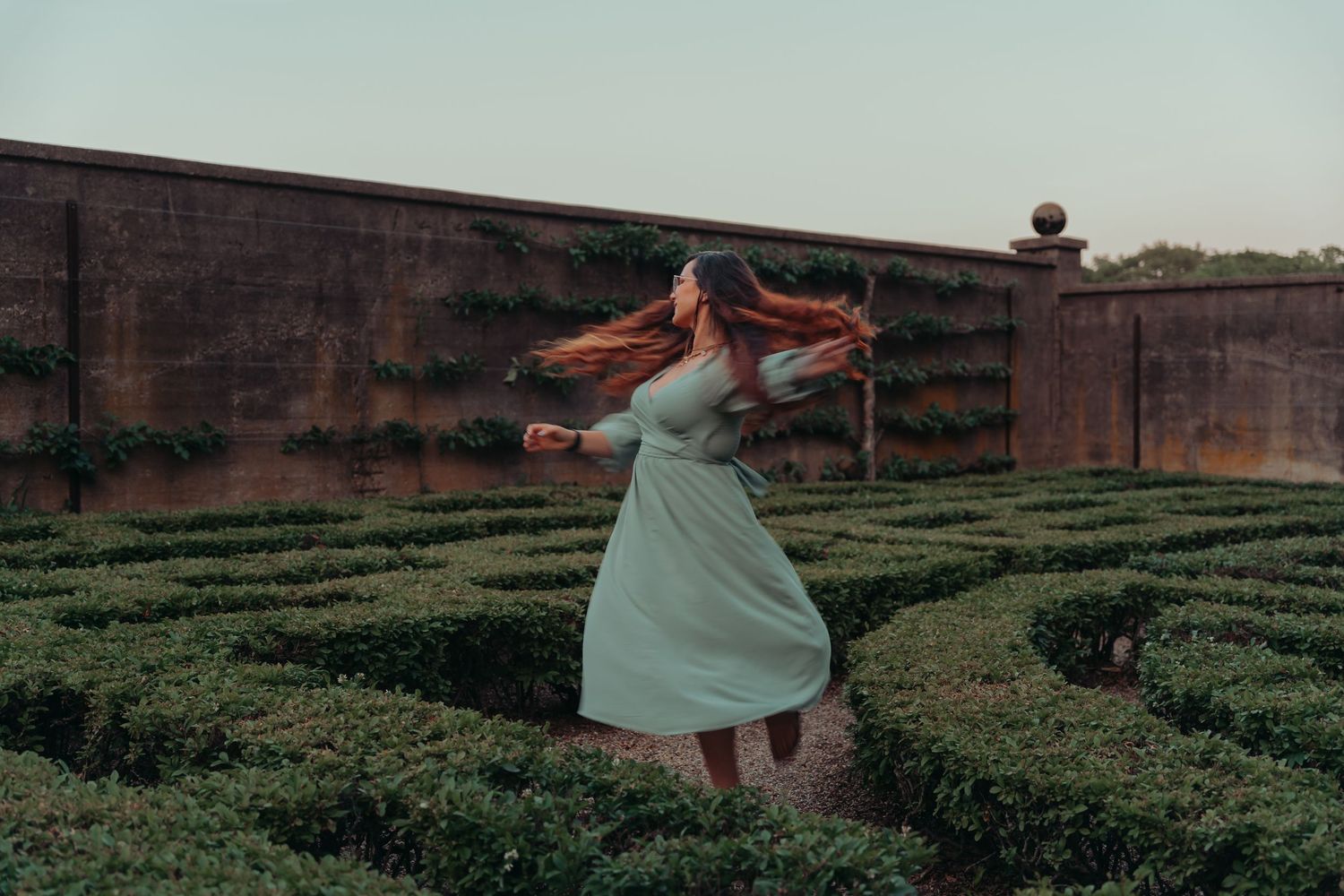 Red-haired woman in a white dress twirling gracefully in a formal hedge maze garden.