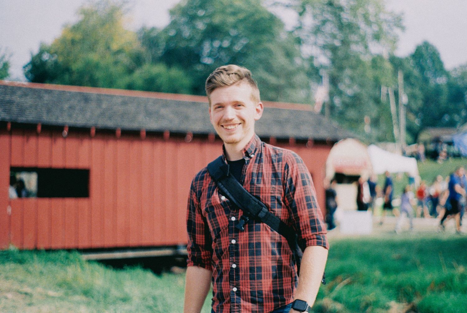 A smiling young man in a plaid shirt stands outdoors near a red barn with horses.