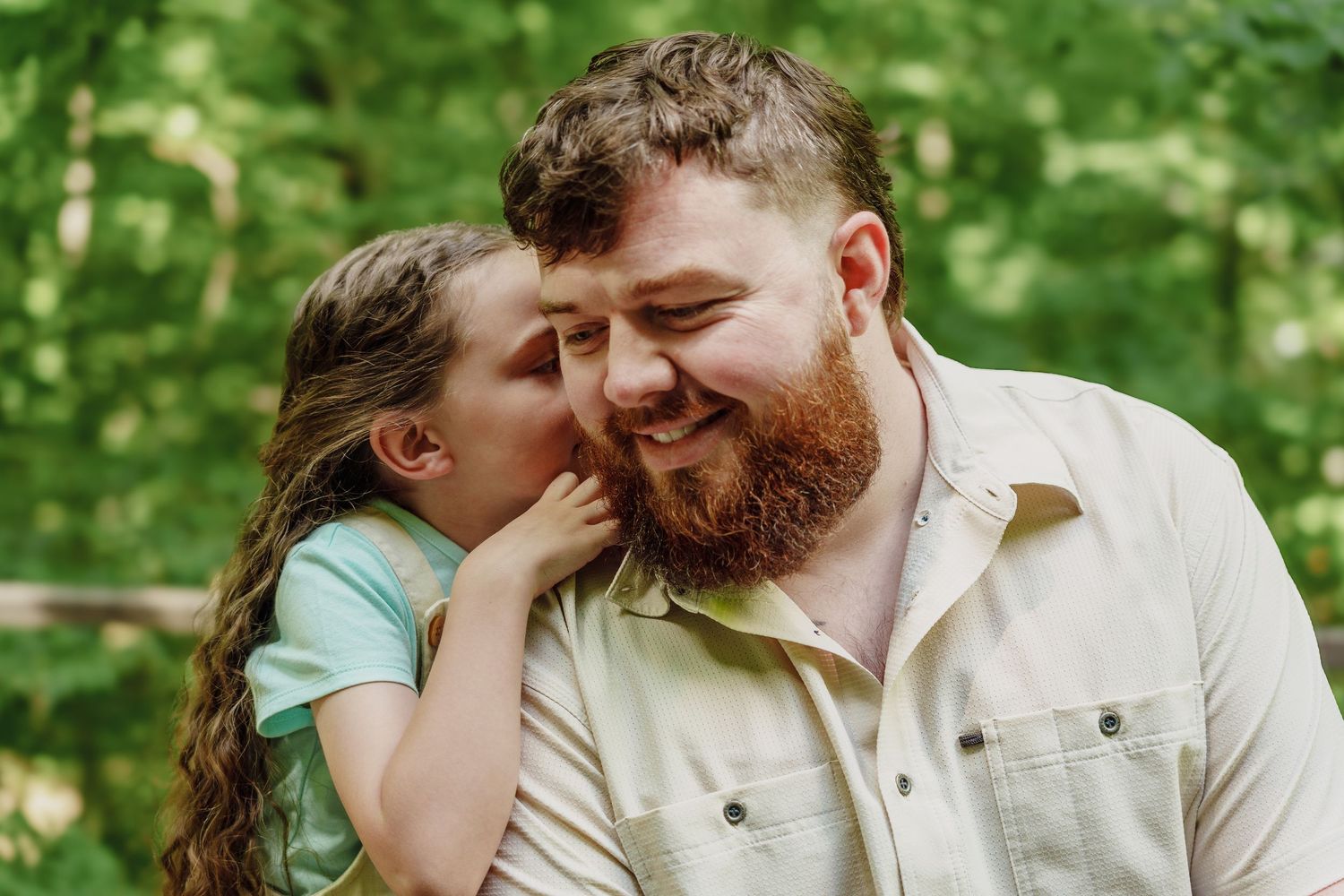 Bearded man with red hair smiling as a young girl whispers in his ear outdoors.