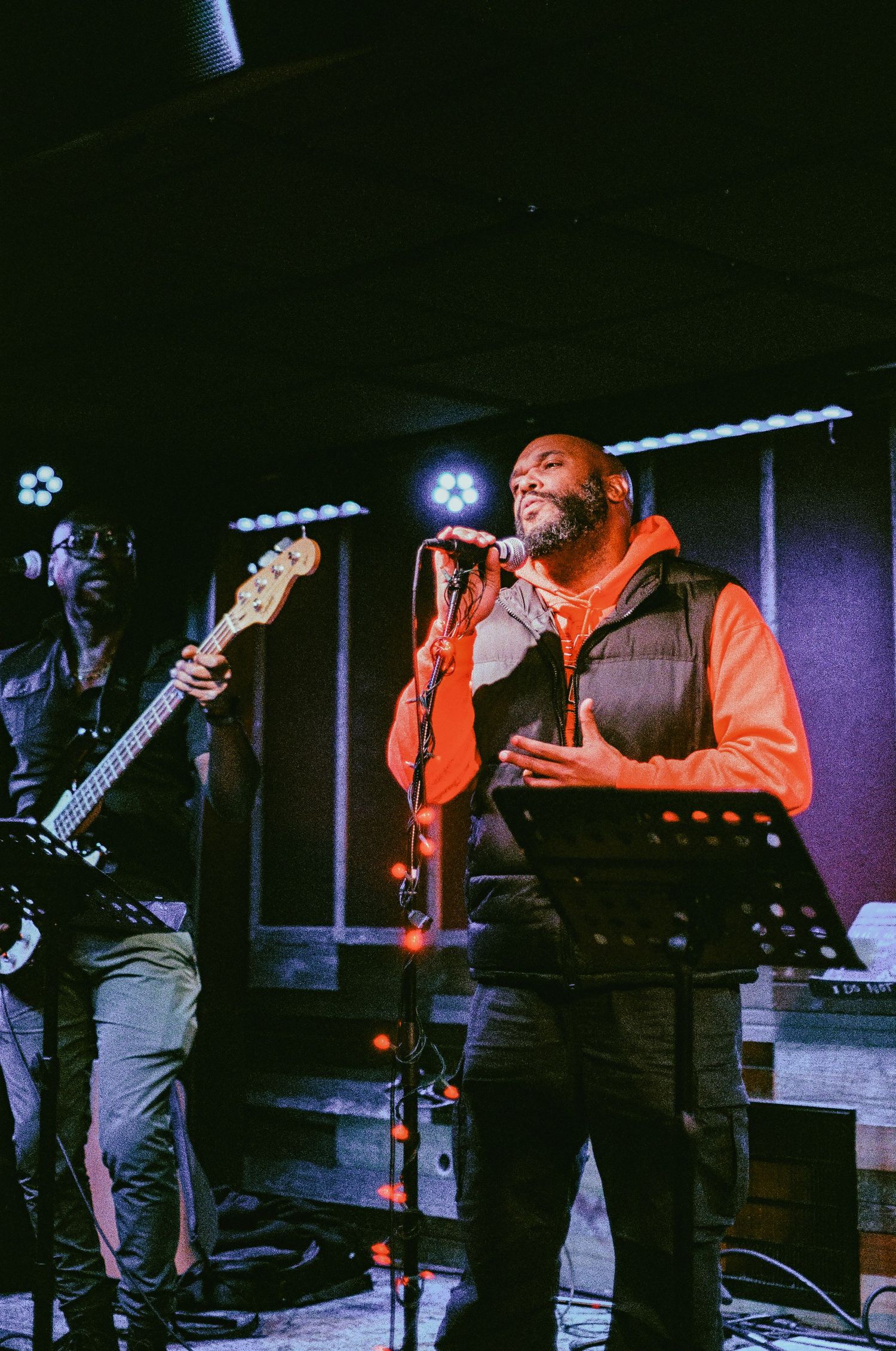 Singer performing live on stage with microphone stand, wearing orange hoodie and black vest, with bassist in background.