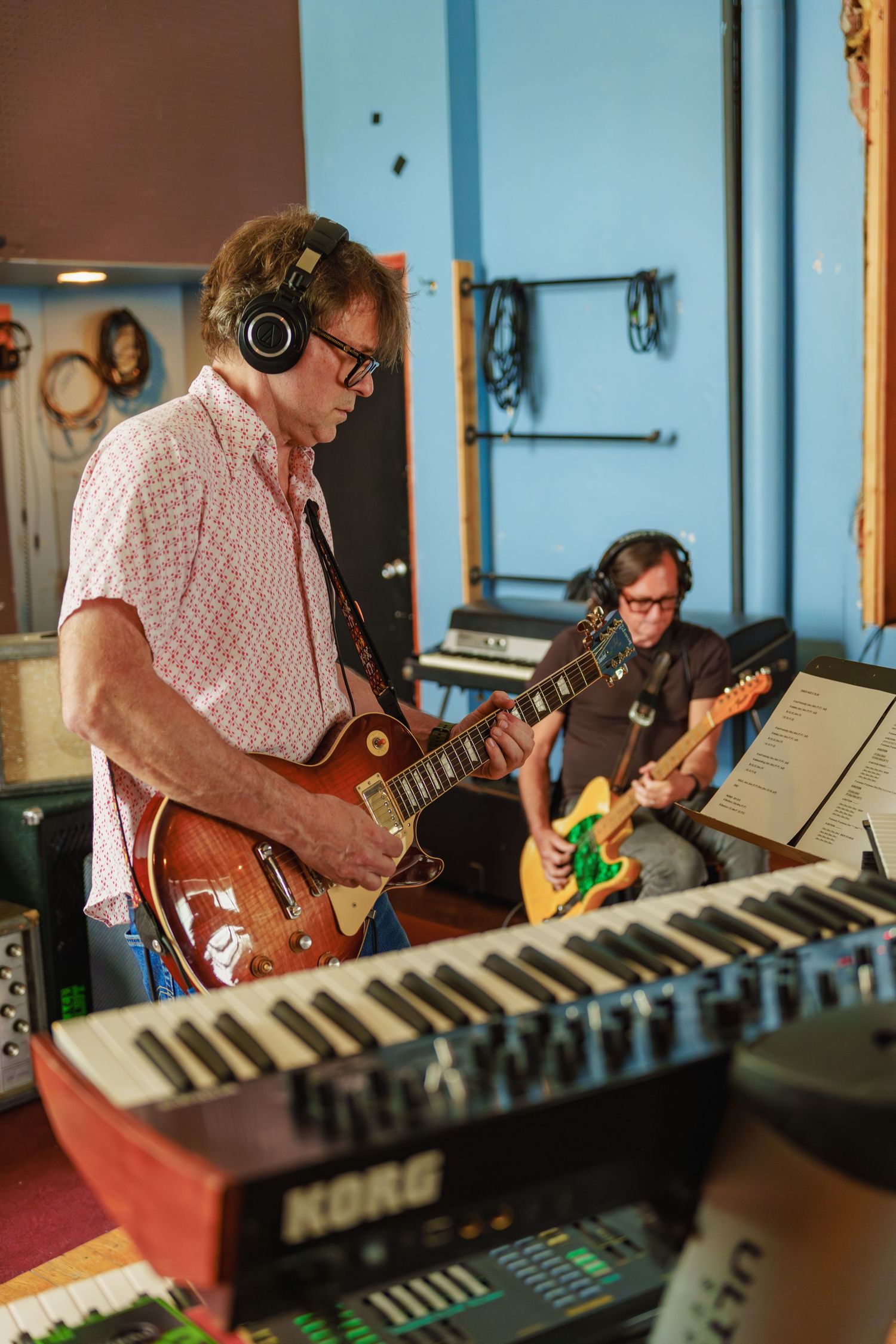 Two guitarists recording in a studio, one wearing headphones playing a Les Paul near a Korg keyboard.