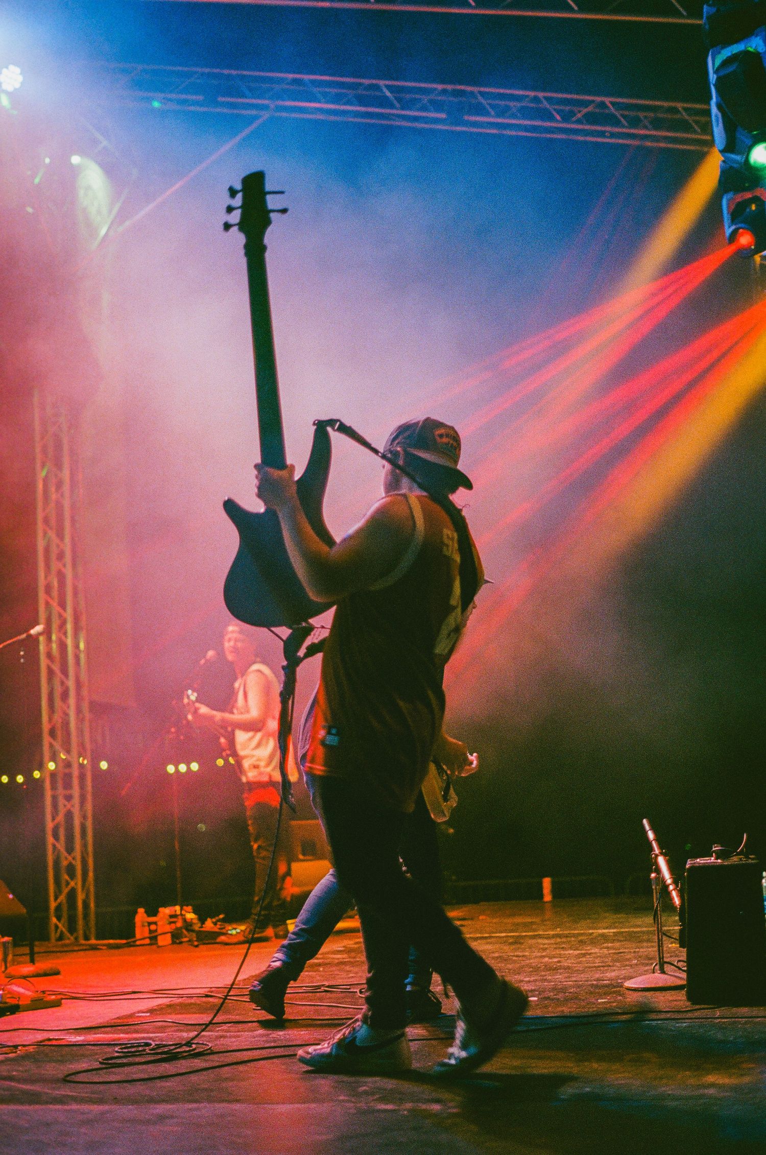Silhouetted bassist holding guitar overhead on a smoke-filled stage with vibrant red and blue lighting.