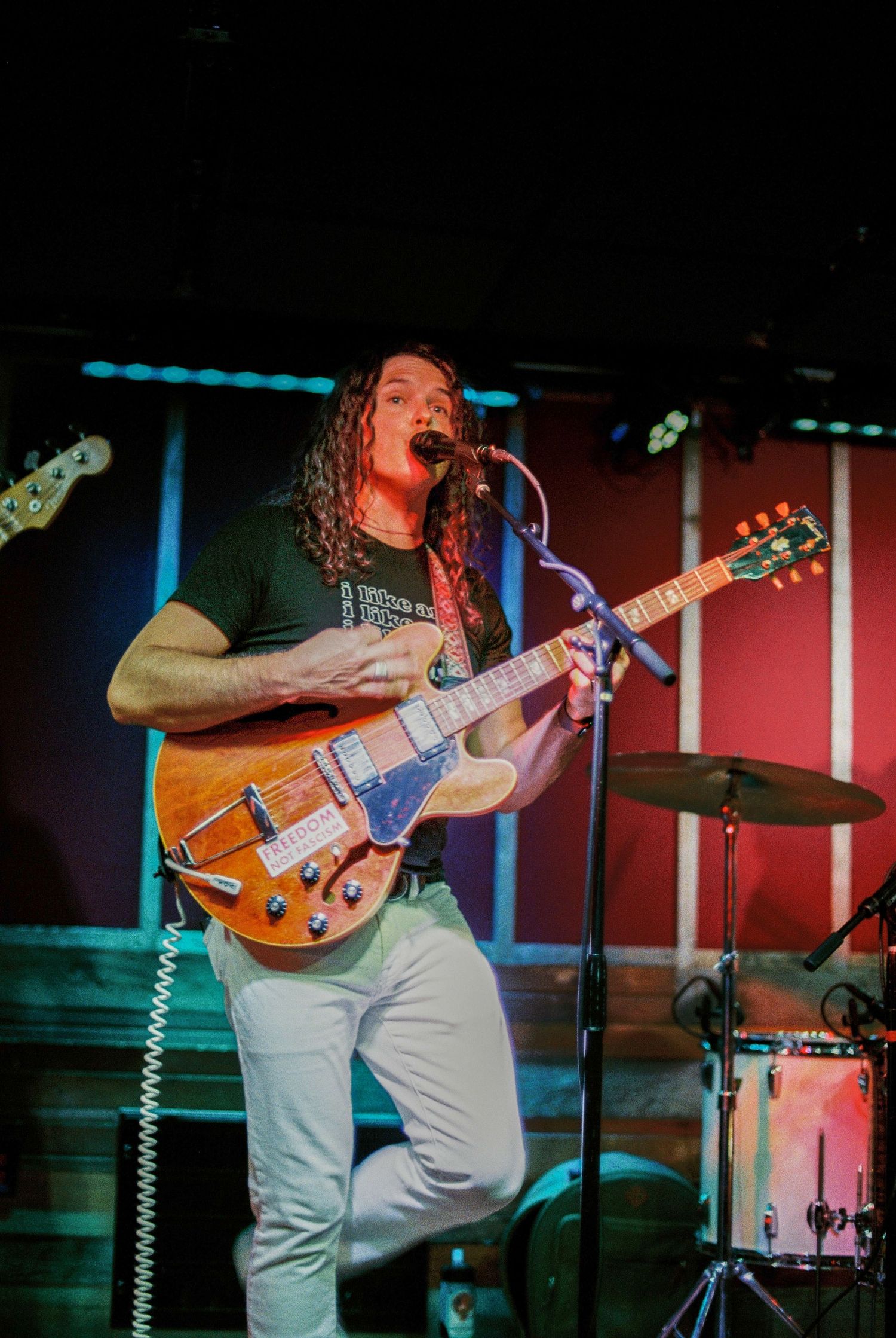 Long-haired musician playing electric guitar and singing on a colorful lit stage.