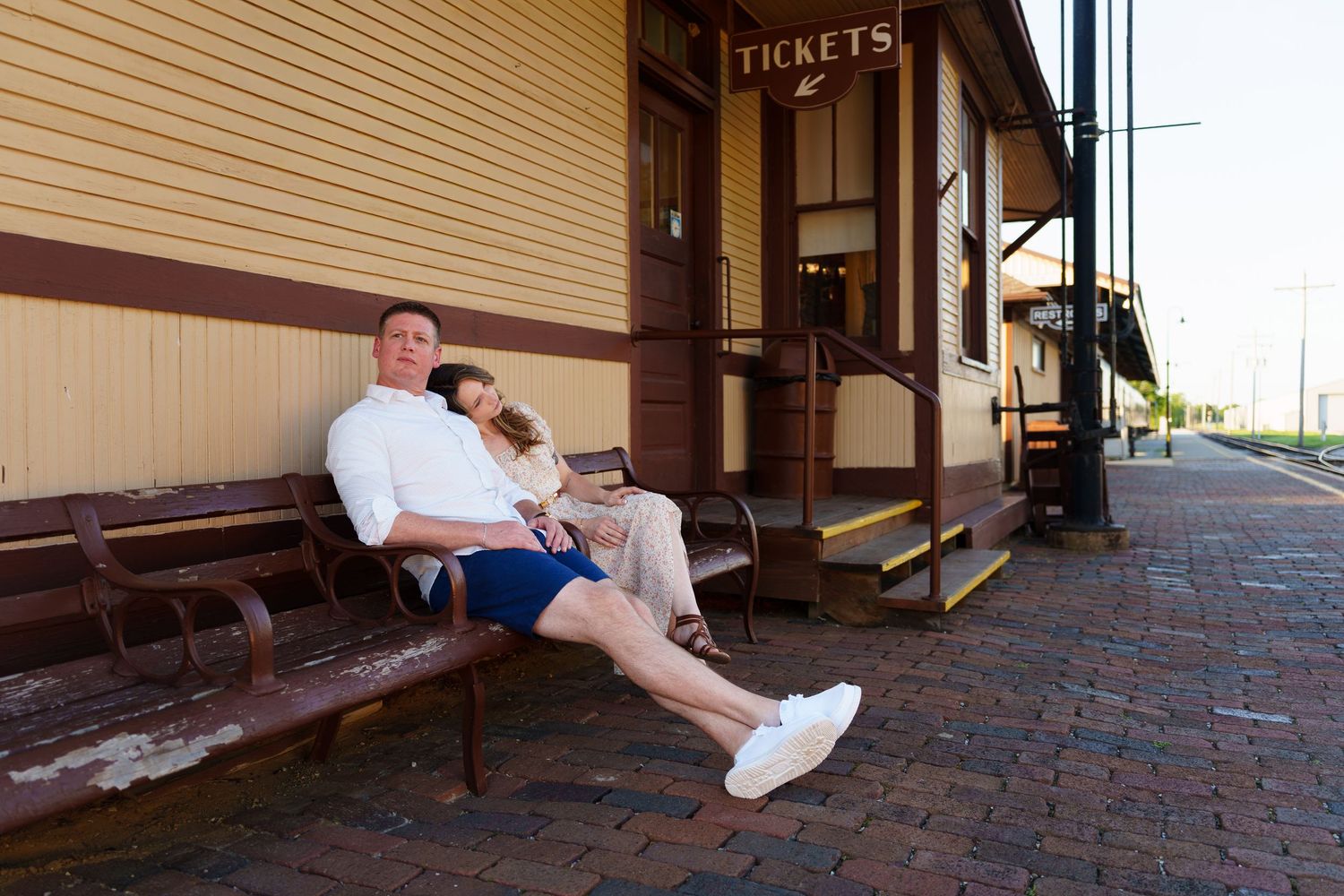 Couple relaxing on a bench outside a vintage train station ticket office on a sunny day.