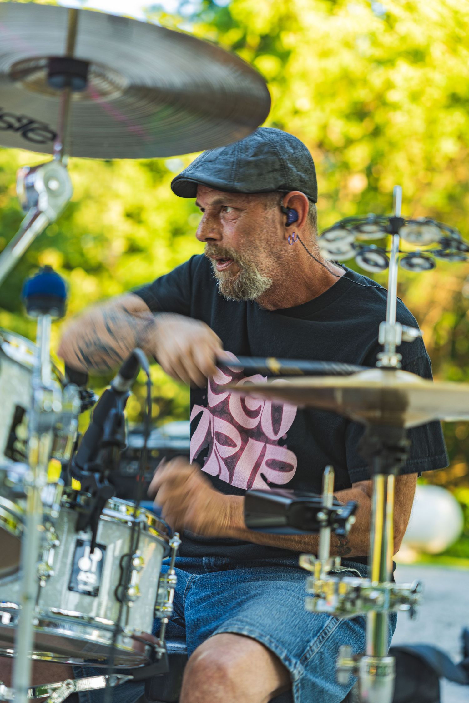 Bearded drummer in black t-shirt and cap playing drums outdoors at a summer music event.