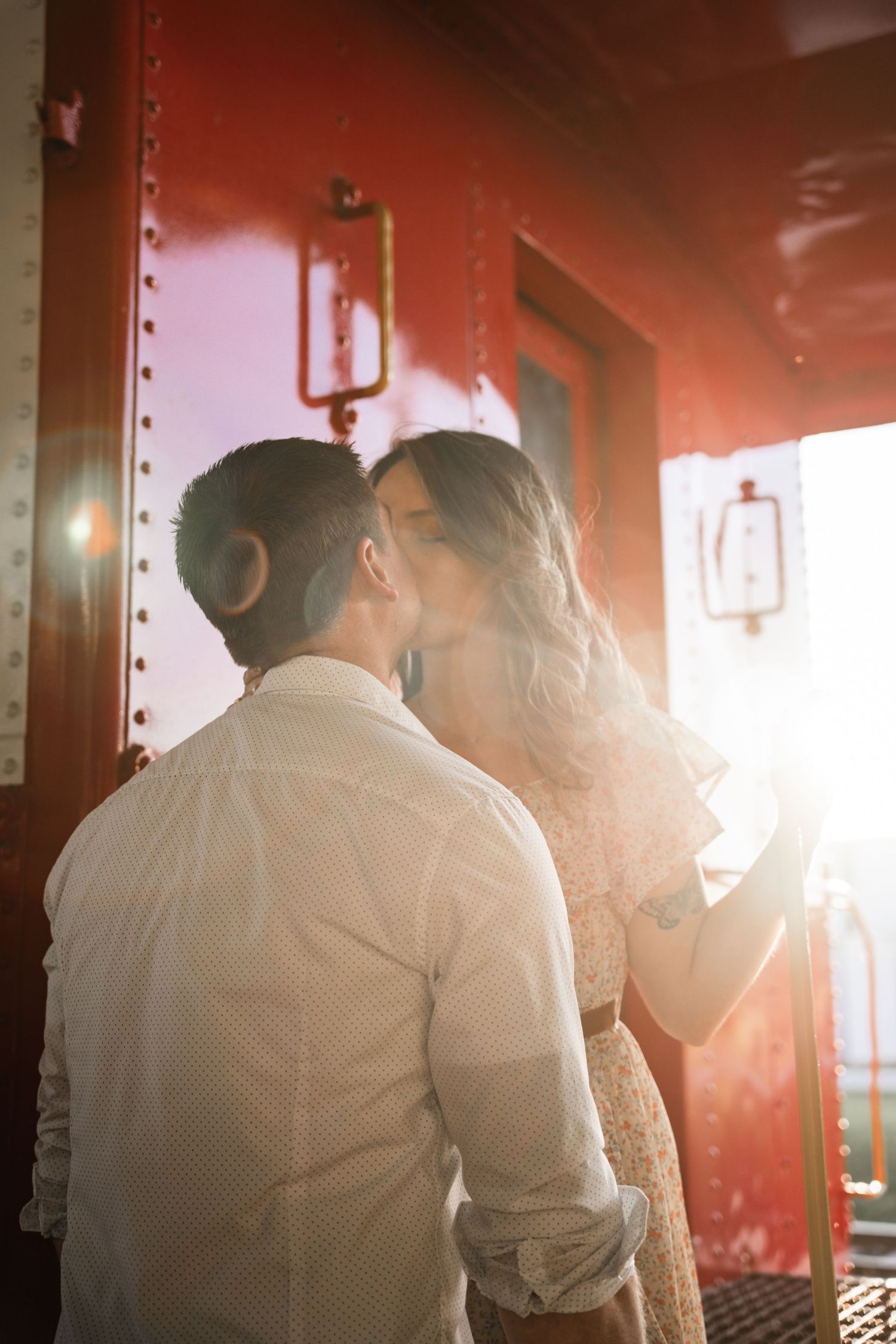 Romantic couple sharing a kiss near a red vintage train car in golden backlit sunlight.