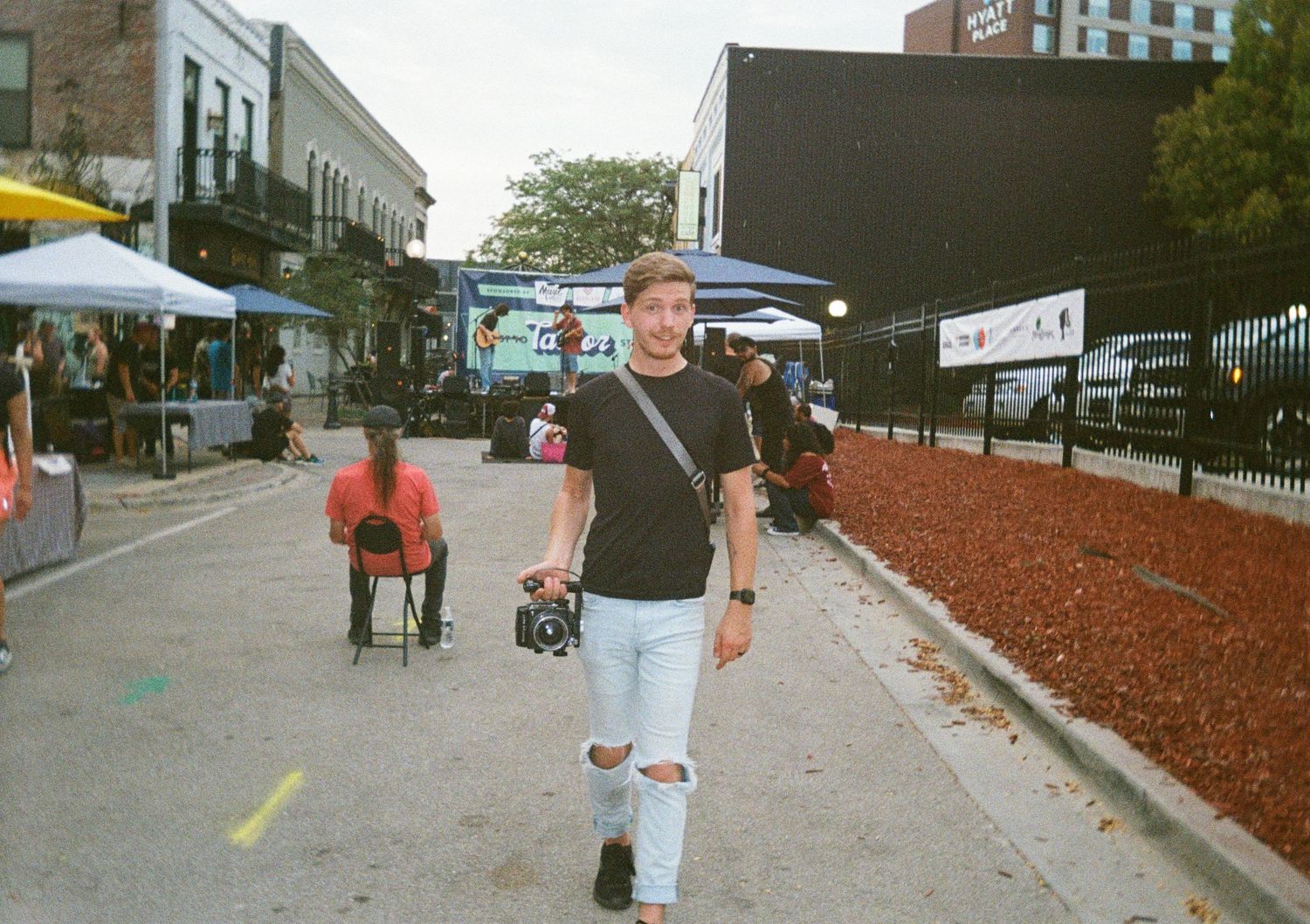 Young man in ripped jeans and black shirt walks down a busy urban street carrying a camera on a sunny day.