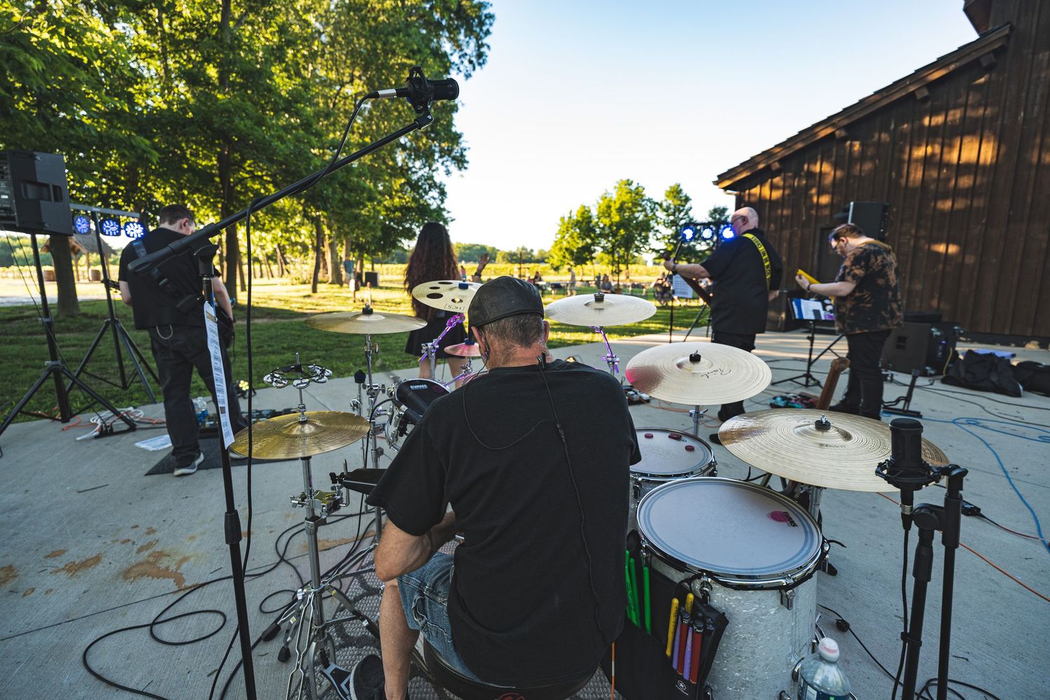 Band performing outdoor concert near rustic barn, viewed from behind the drummer on a sunny day.