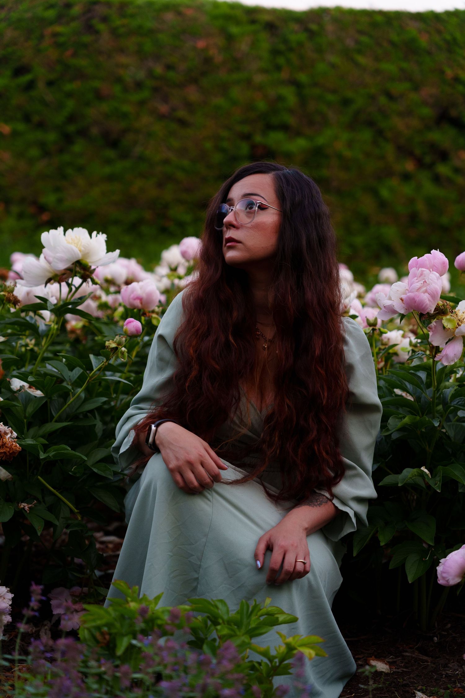 Woman with long red hair and glasses sitting among blooming pink and white peonies.