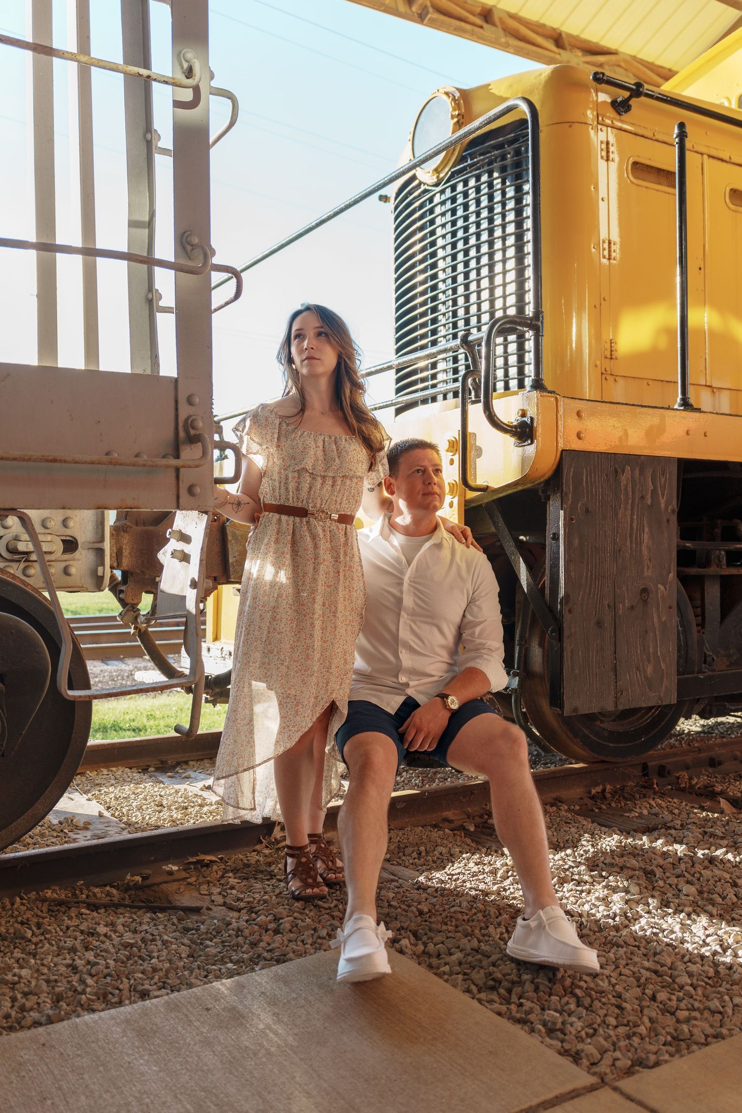 Couple posing beside a yellow train locomotive on sunny railroad tracks outdoors.
