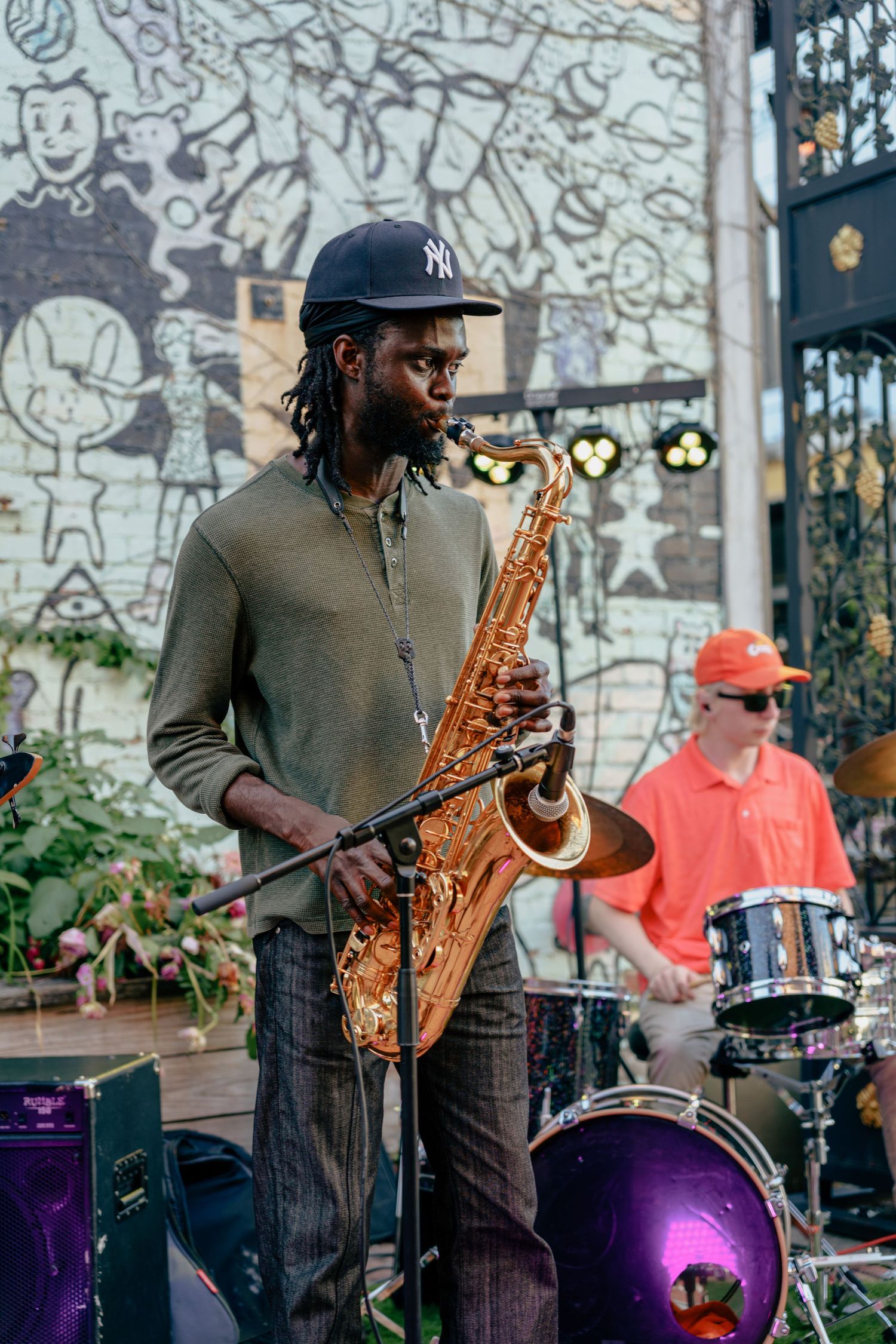 A jazz musician plays saxophone outdoors at a street performance with a colorful mural backdrop.